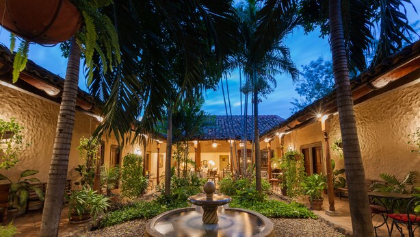 A courtyard with a central fountain, surrounded by palm trees and potted plants, leading to a building with a tiled roof and warm lighting. - Los Robles Charming Colonial Boutique Hotel (Managua)