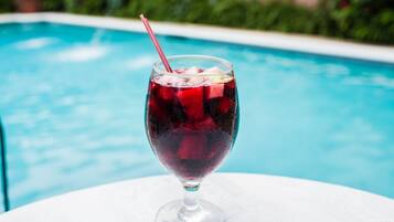 A glass of red drink with ice cubes on a white table by a pool.