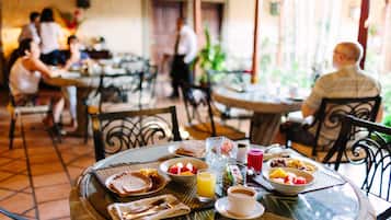 A dining area with round tables, chairs, and a variety of breakfast items including bread, fruit, and beverages.