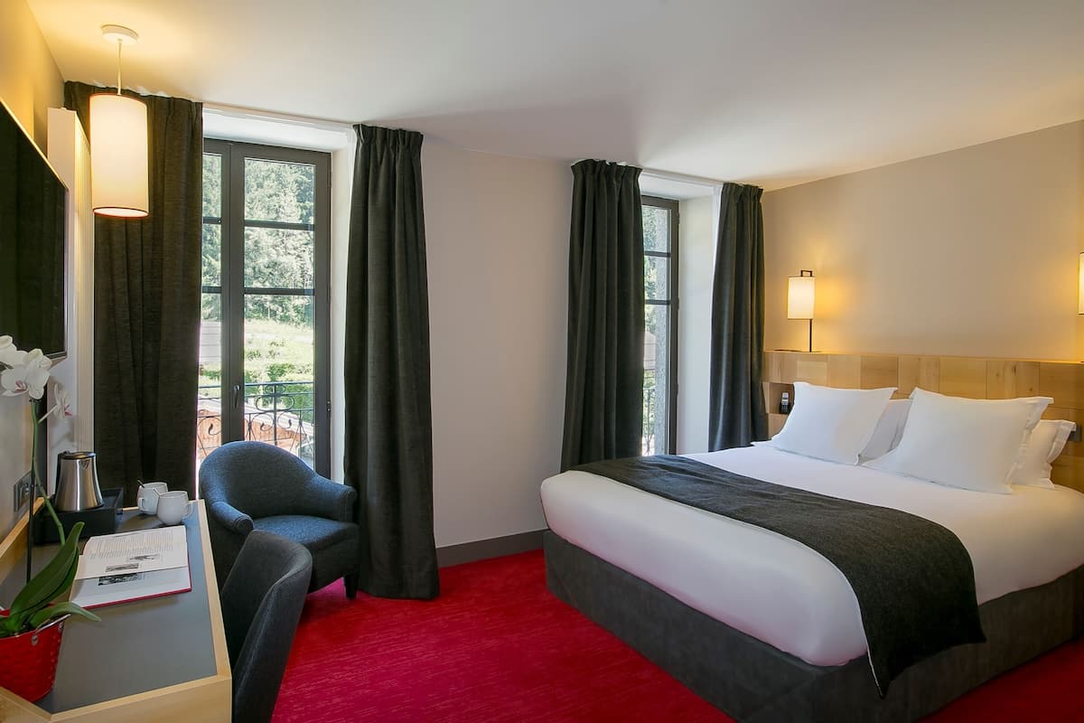 A hotel room with a large bed, a desk with a chair, a red carpet, and a view of greenery through the windows.