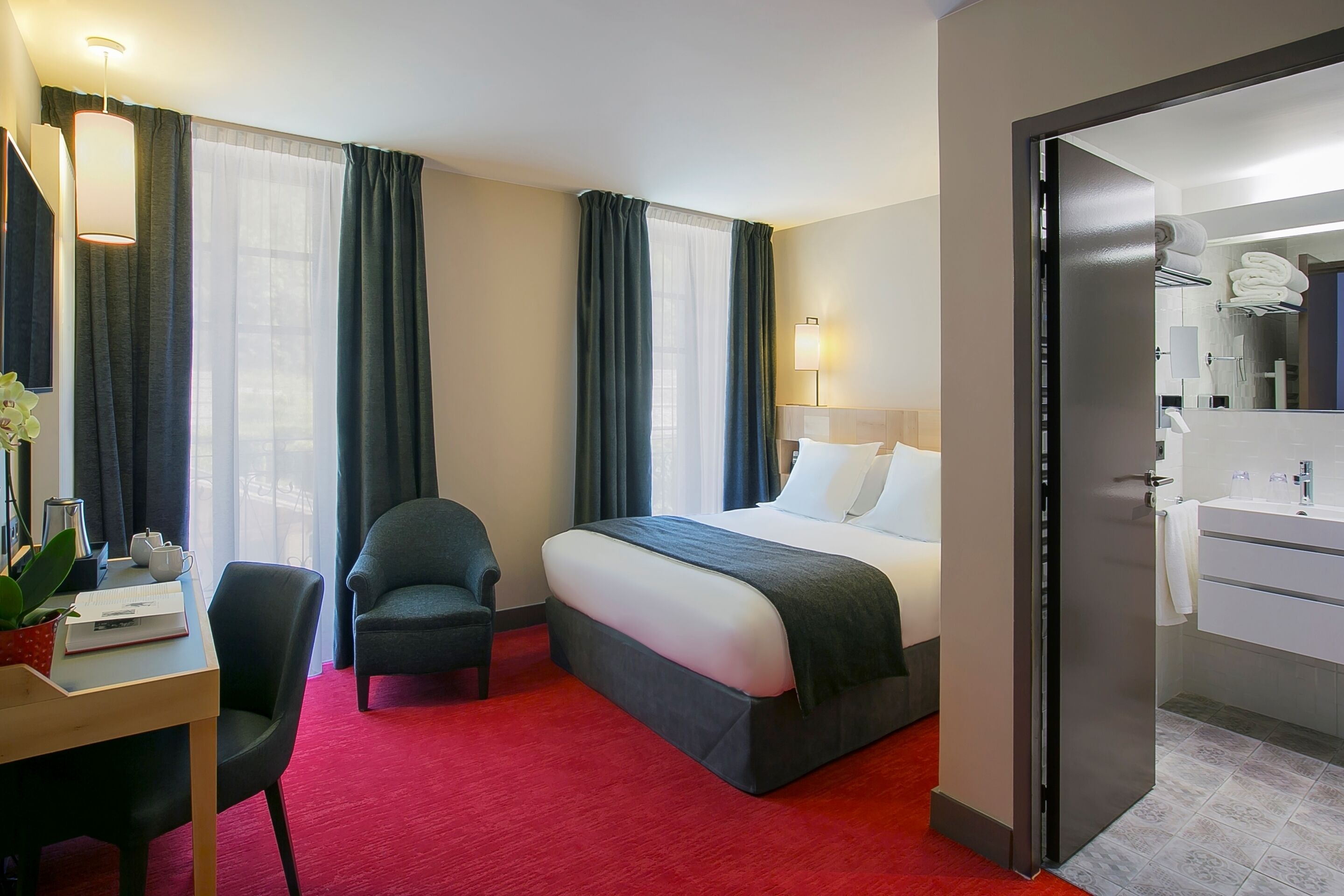 Ein Hotelzimmer mit einem Bett, einem Schreibtisch mit Stuhl, einem kleinen Tisch mit Pflanze und einem durch eine offene Tür einsehbaren Badezimmer.