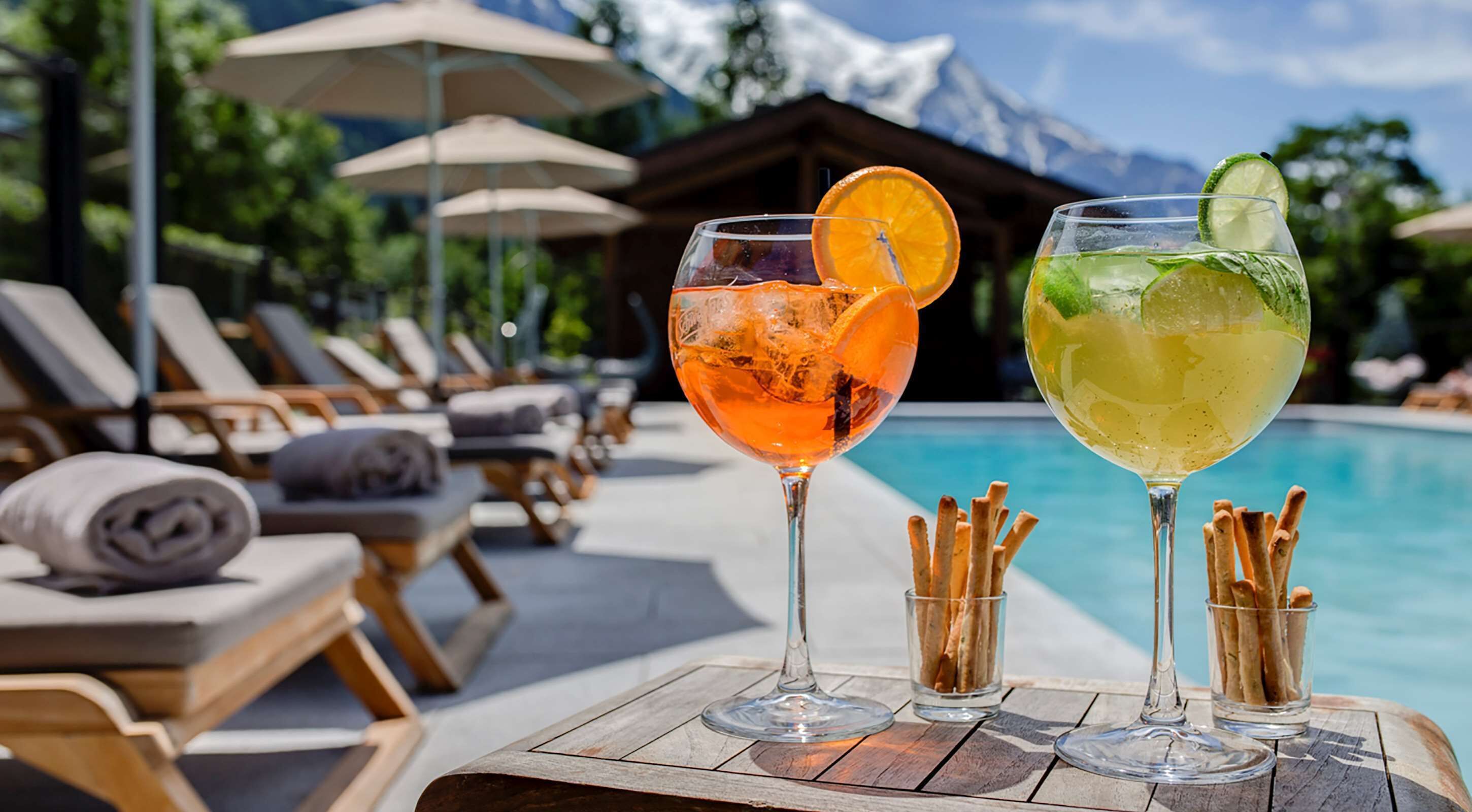 To cocktails på et træbord ved en pool med liggestole og parasoller i baggrunden.