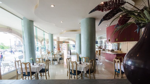 A spacious indoor dining area with multiple tables and chairs, large windows, and a central decorative plant. - San Francisco Business Class (Irapuato)