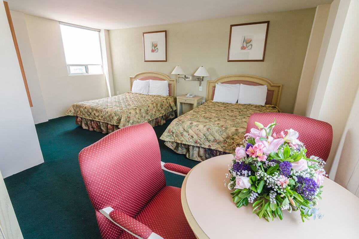 A hotel room with two beds, a red chair, a small table with a floral arrangement, and a window providing natural light.