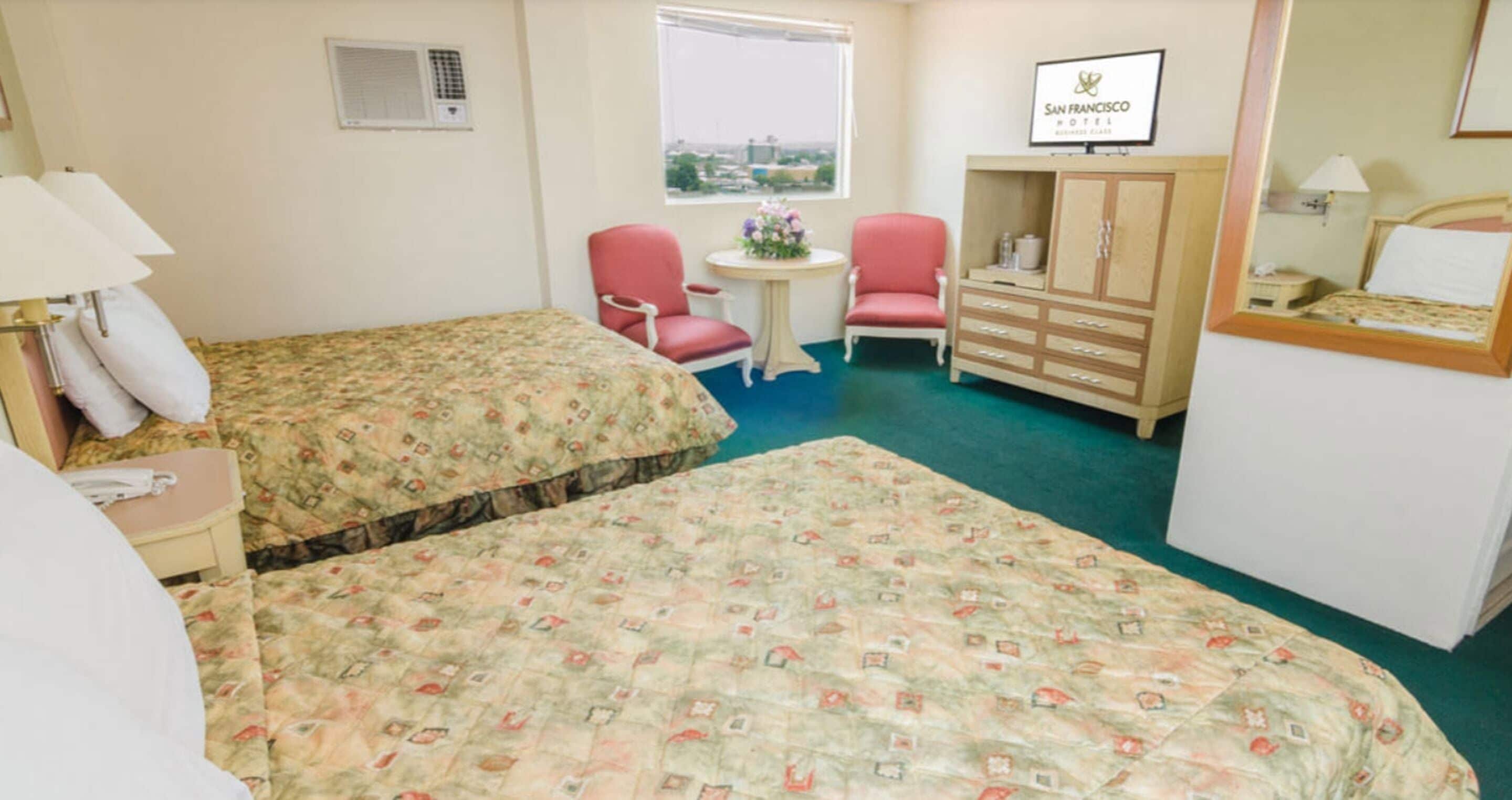 Habitación de hotel con dos camas, un escritorio, una silla, una mesita con flores, un armario y una ventana con vista a la ciudad.