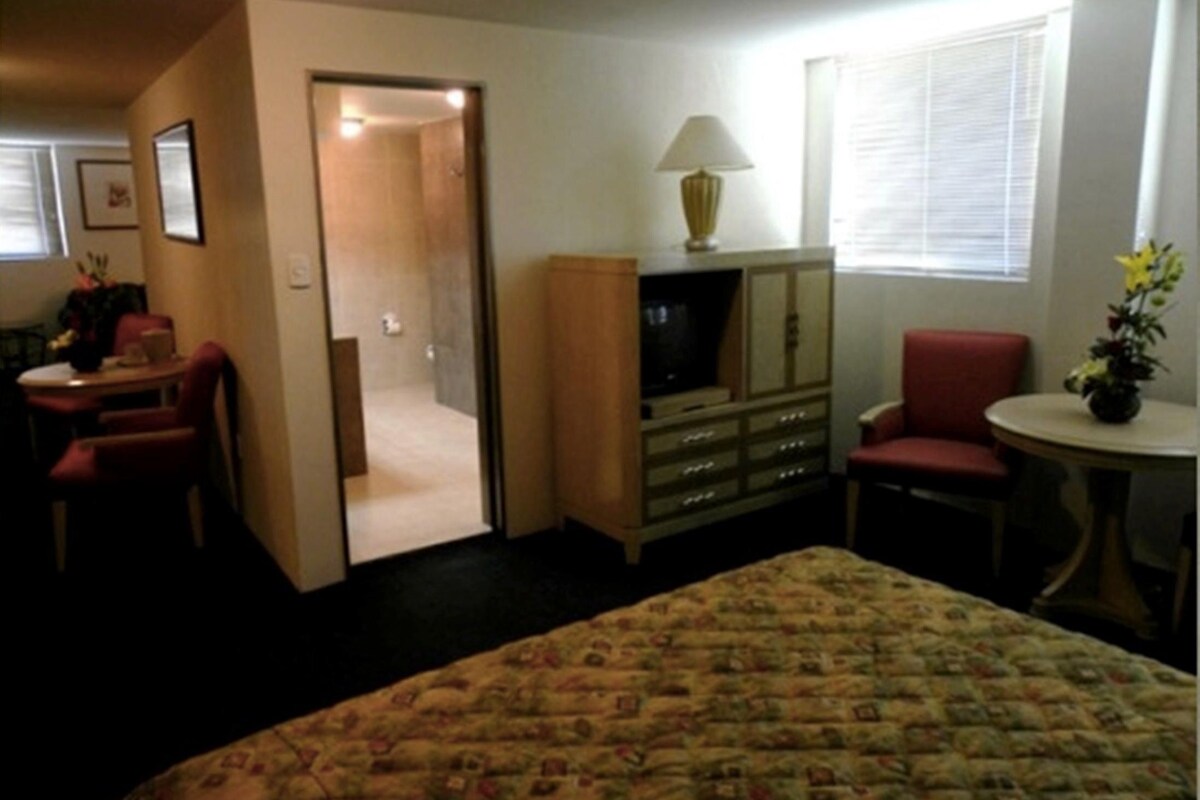 Habitación de hotel con cama, escritorio, silla, mueble para televisión y ventana con persianas.