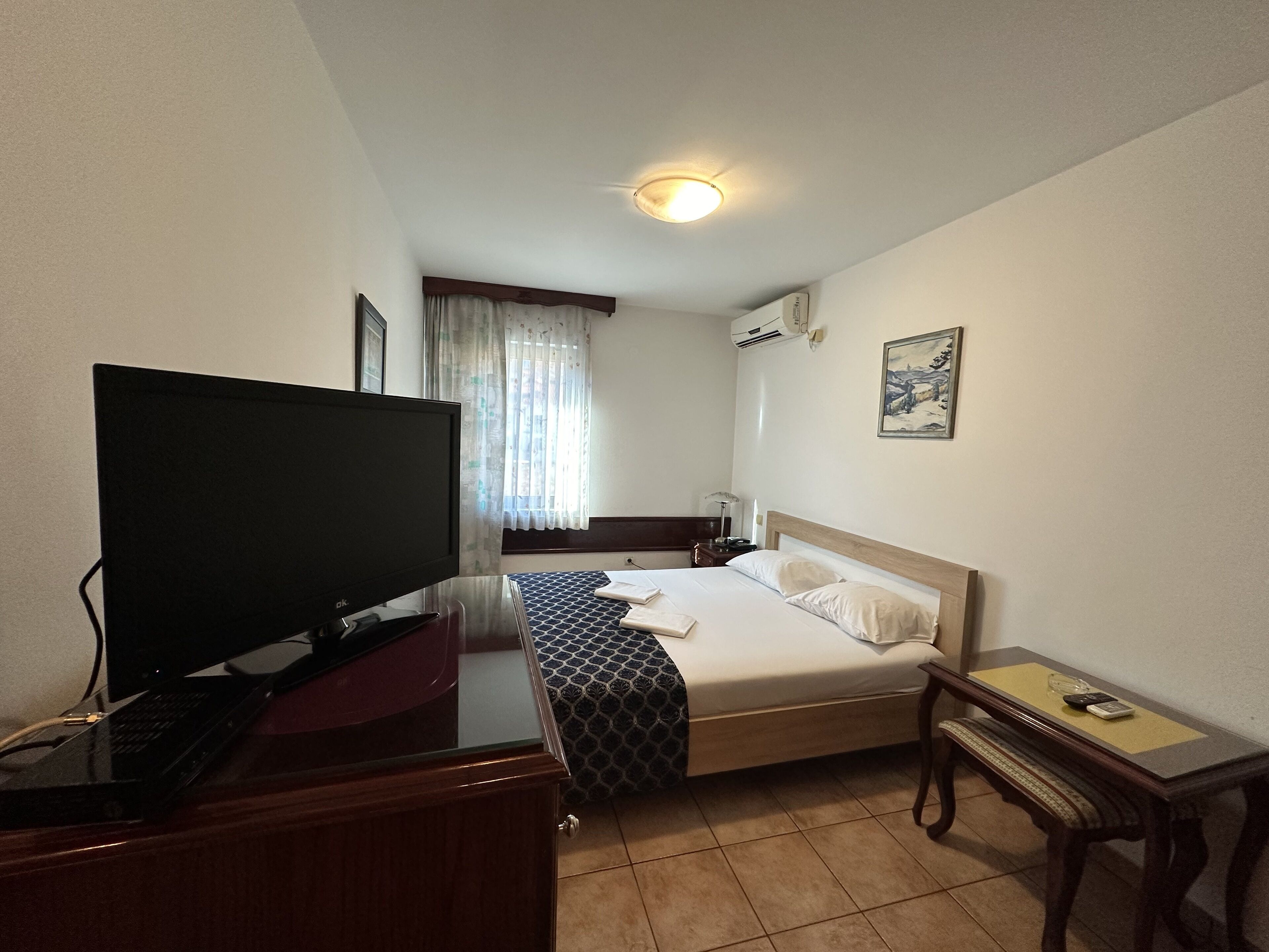 A hotel room with a bed, a desk with a computer, a TV, a bedside table, and a window with curtains.