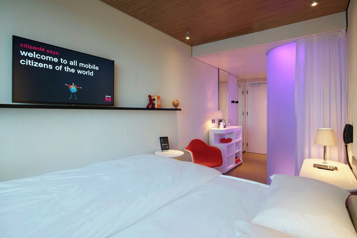 A modern hotel room with a bed, a red chair, a small table, and a TV displaying a welcome message.