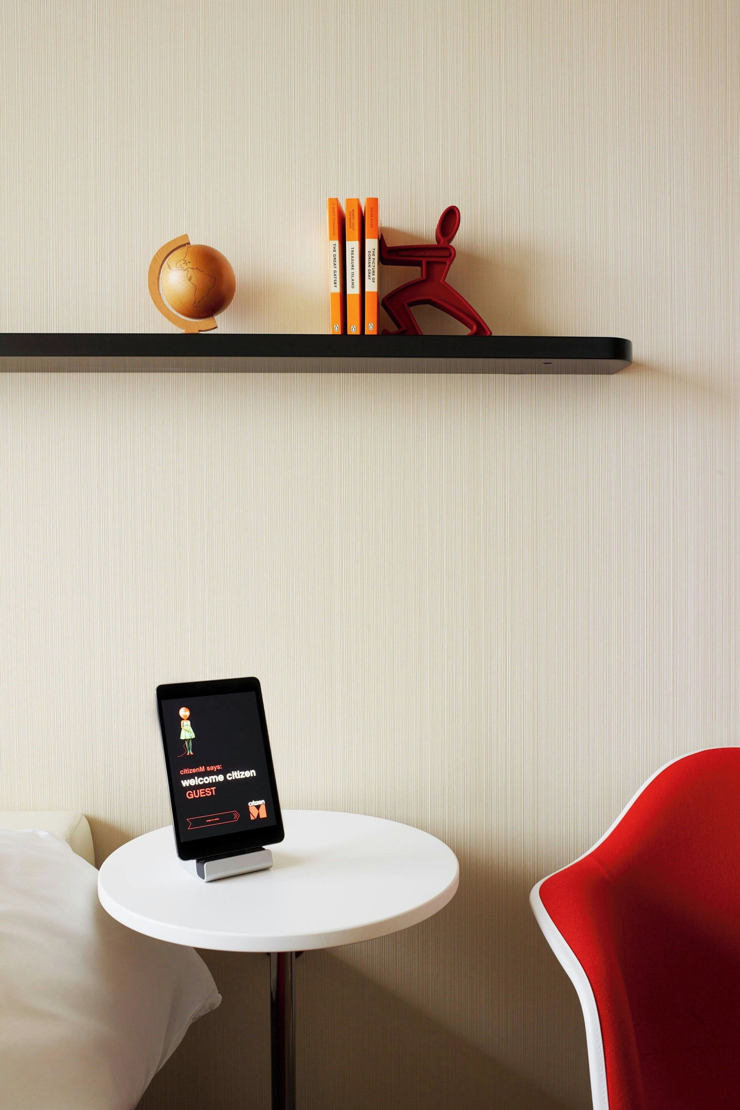 Habitación de hotel con una mesa redonda blanca, una silla roja, una estantería con libros y un objeto decorativo, además de una tableta mostrando un mensaje de bienvenida.