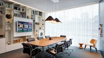 Uma sala de conferências moderna com uma grande mesa de madeira, cadeiras de escritório pretas, uma televisão na parede e uma ampla janela com persianas.