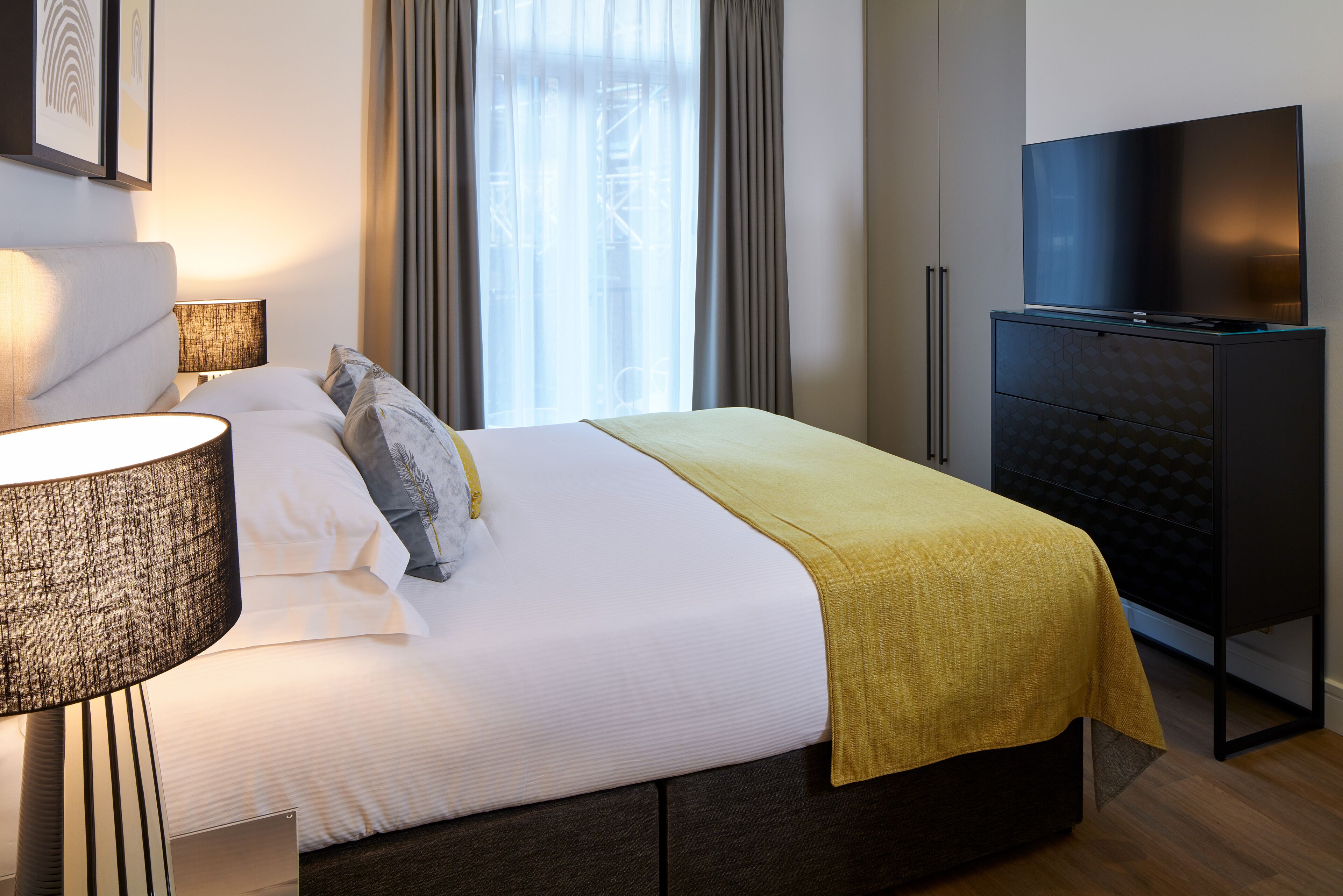 A neatly made bed with a white comforter and a yellow throw blanket, a bedside lamp, and a flat-screen TV on a stand.