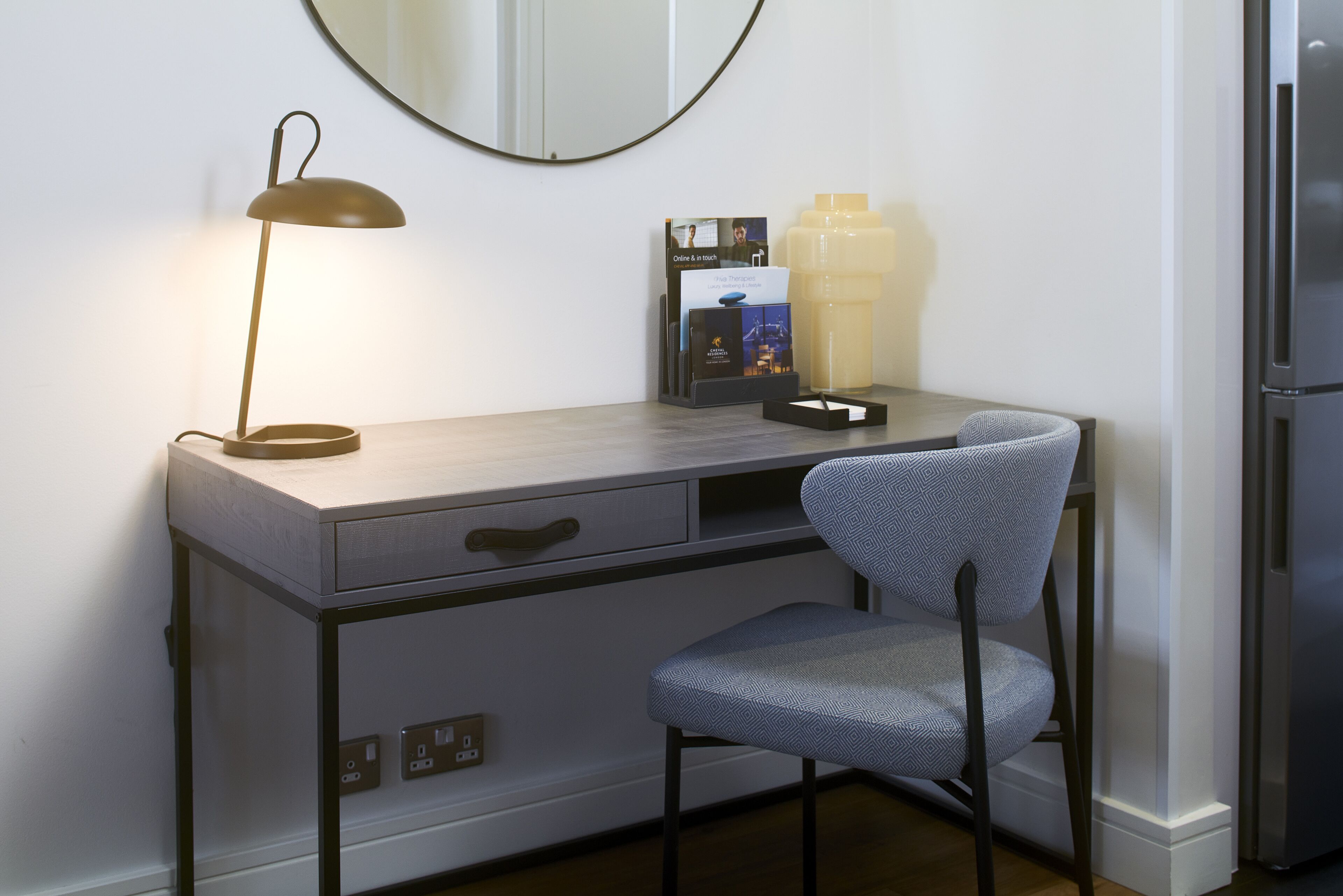 A desk with a lamp, a chair, a large water jug, and a refrigerator.