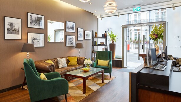 Una hall con un divano, poltrone, un tavolino da caffè, quadri incorniciati alle pareti, una reception e un'ampia finestra con vista sugli edifici.