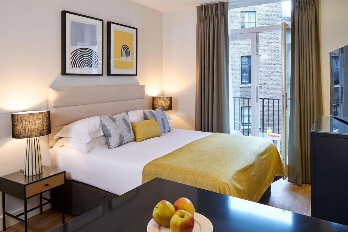 A hotel room with a bed, bedside lamps, a desk with apples, and artwork on the wall.