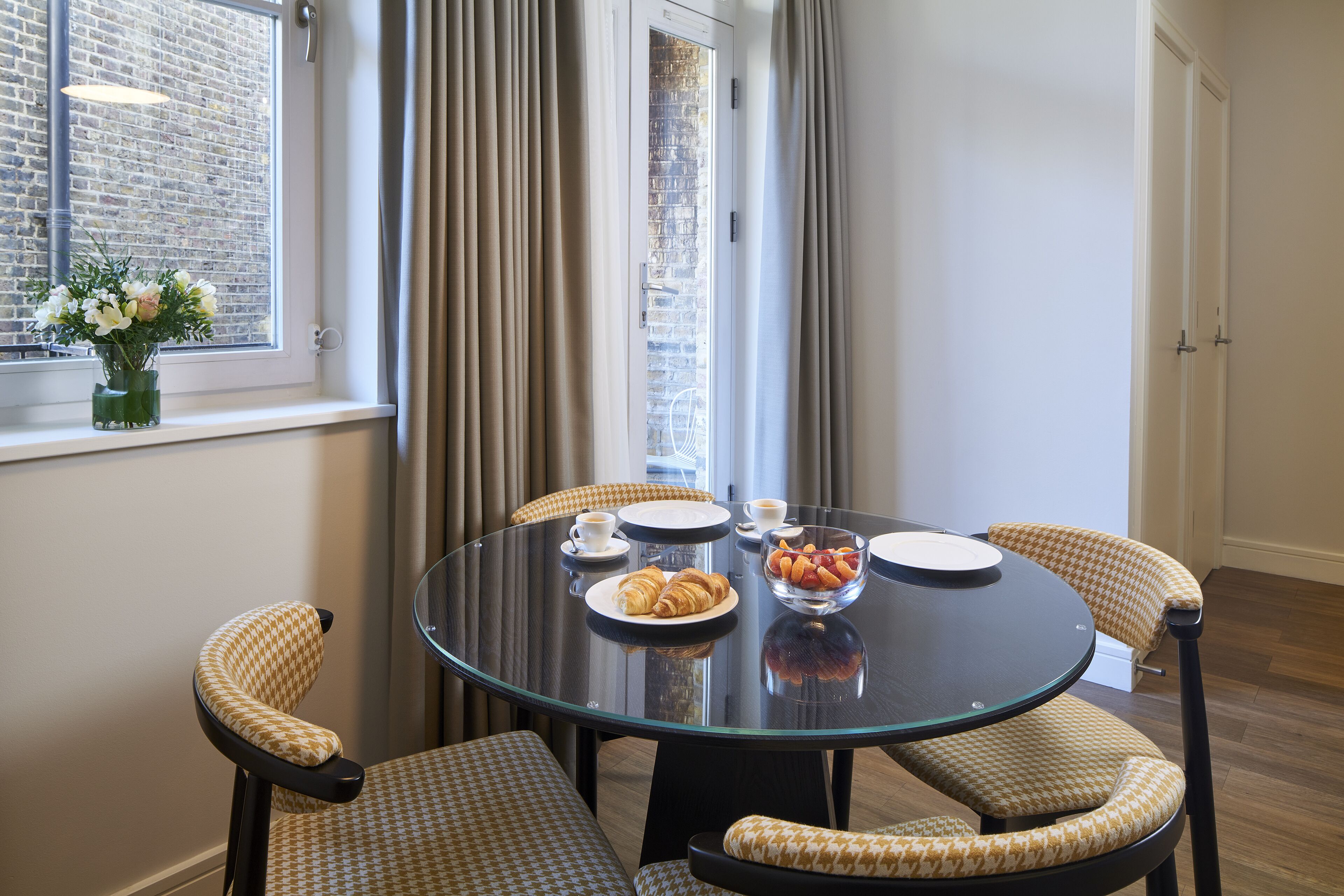 A dining area with a round glass table, checkered chairs, and a vase of flowers on the windowsill.