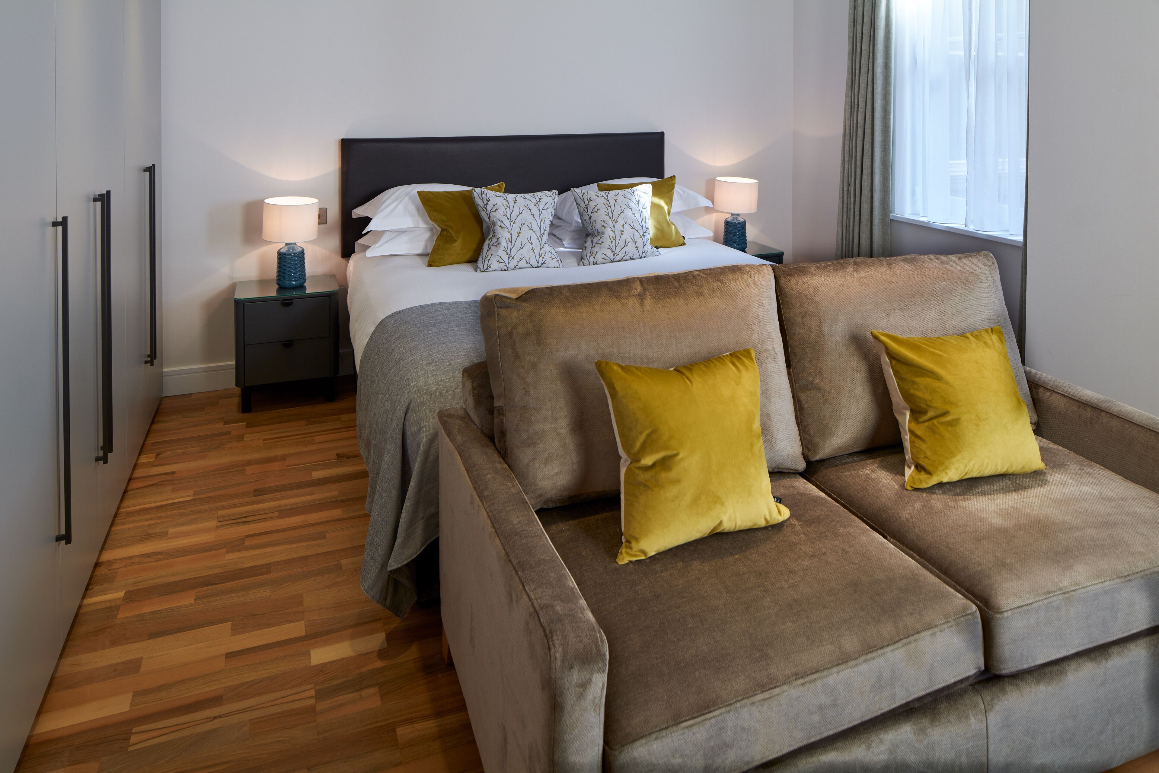 A modern hotel room with a bed, two nightstands, a sofa, and a wooden floor.