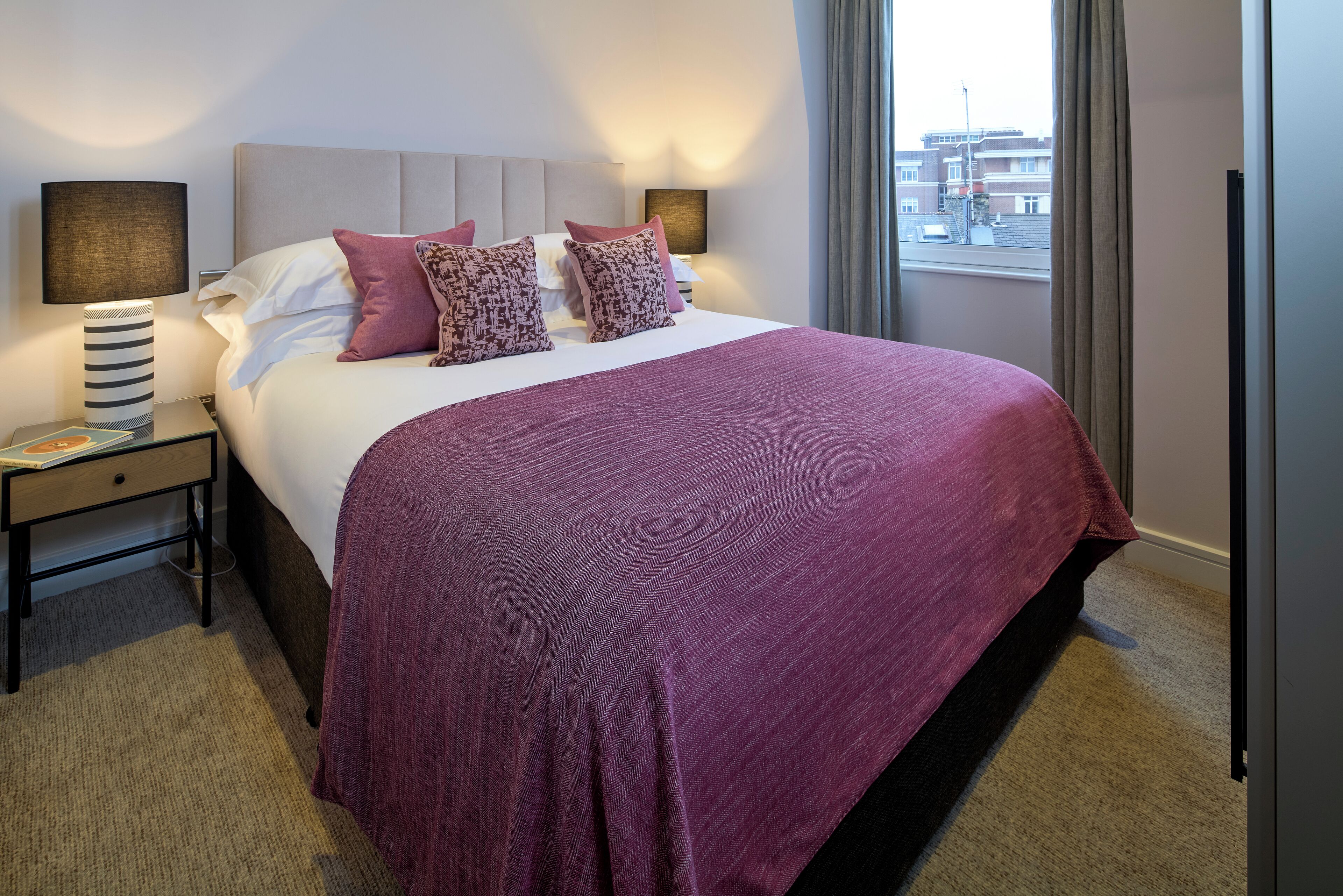 A hotel room with a large bed, a nightstand with a lamp, and a view of the city through the window.