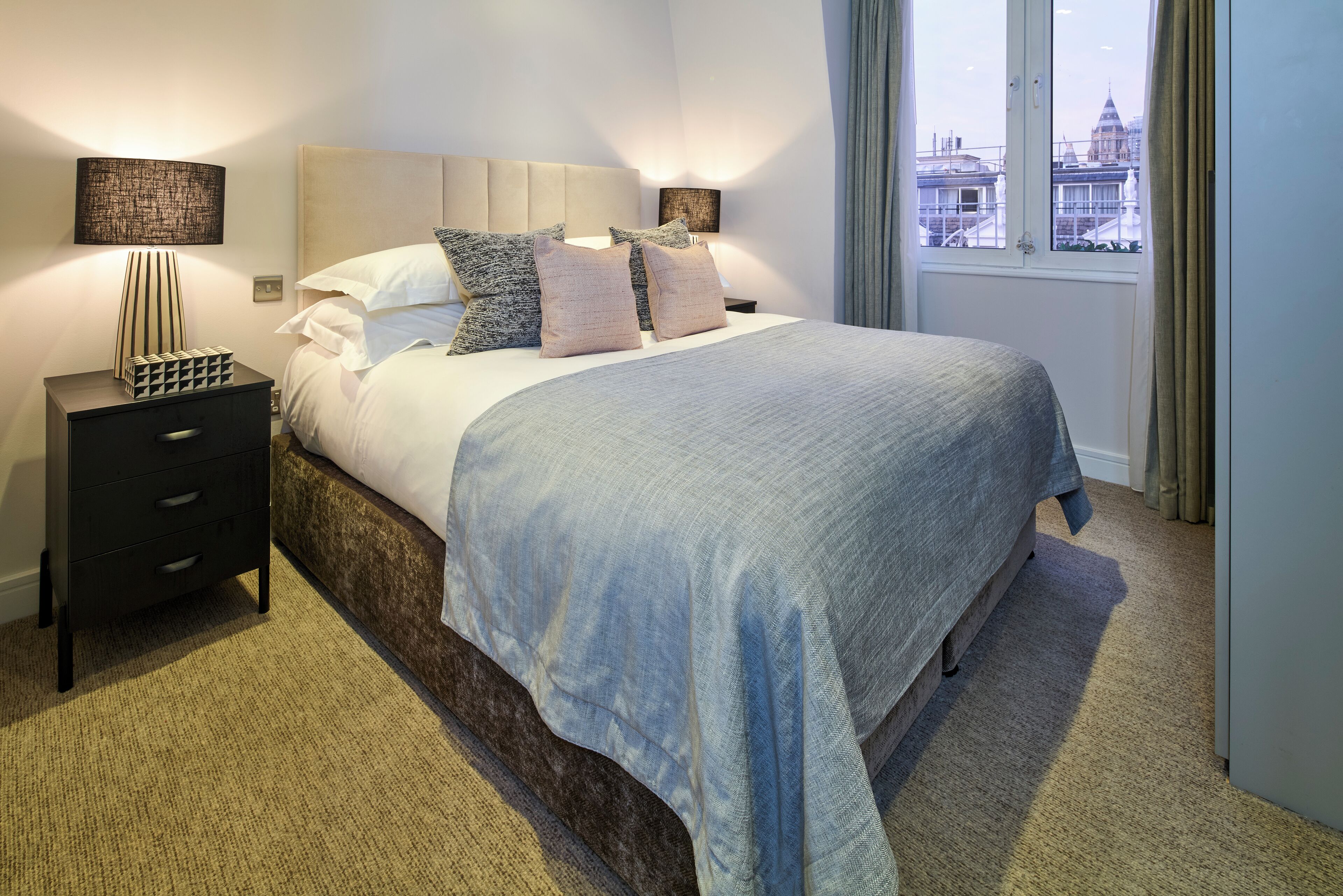 A hotel room with a large bed, a nightstand, a lamp, and a view of a cityscape through a window.