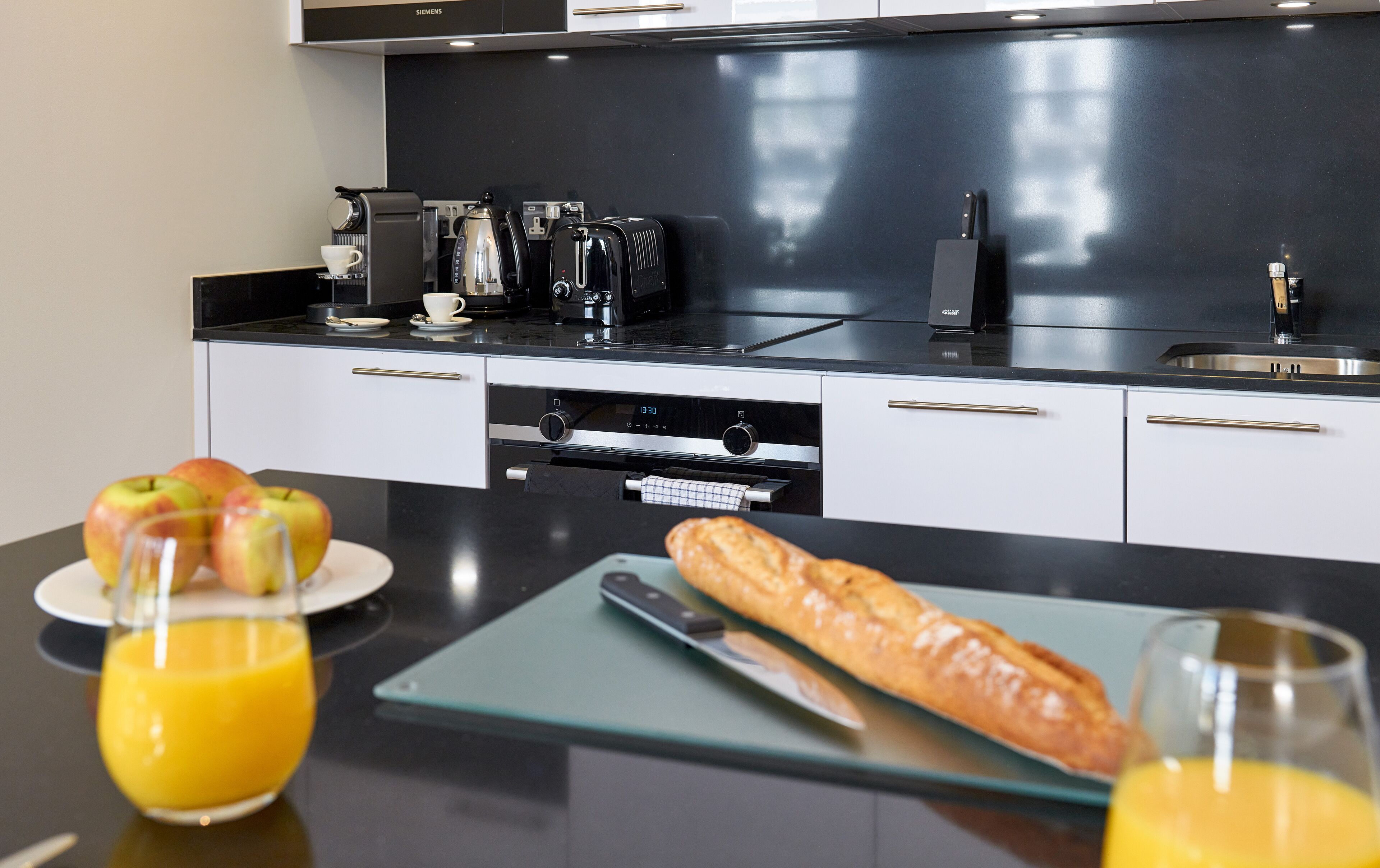 A modern kitchen with a built-in coffee maker, toaster, and sink. There are apples and a baguette on the counter, and a glass of orange juice.