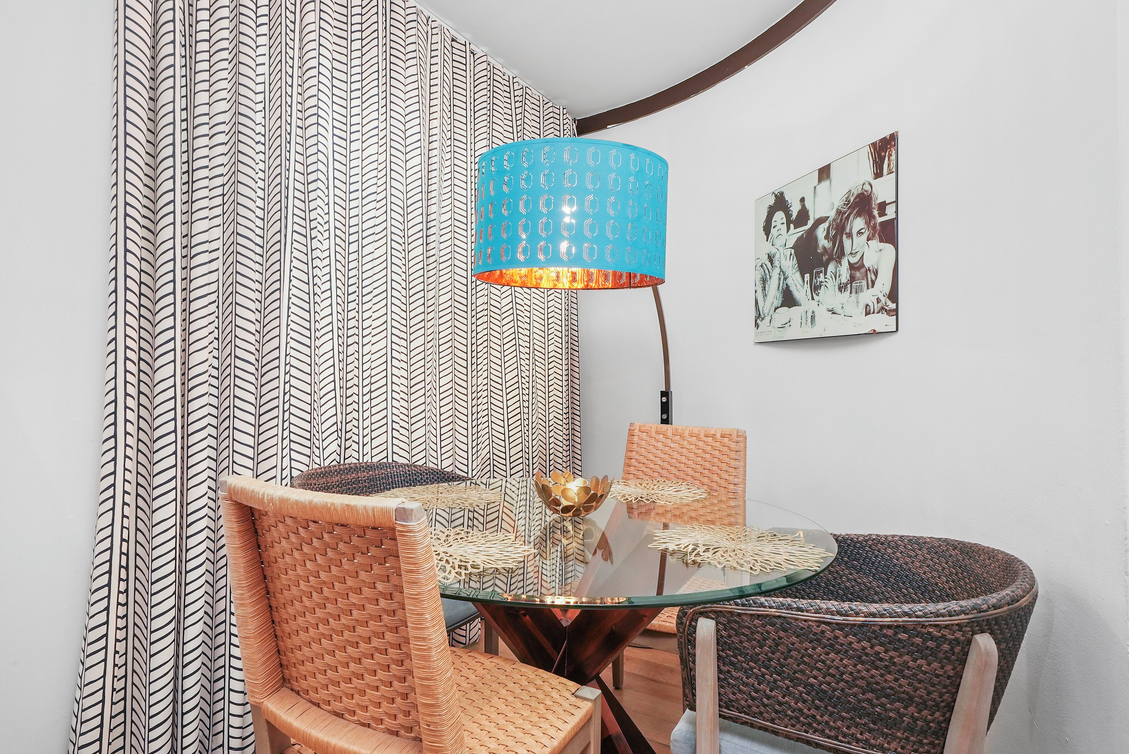 A dining area with a glass table, wicker chairs, a blue lamp, and patterned curtains.