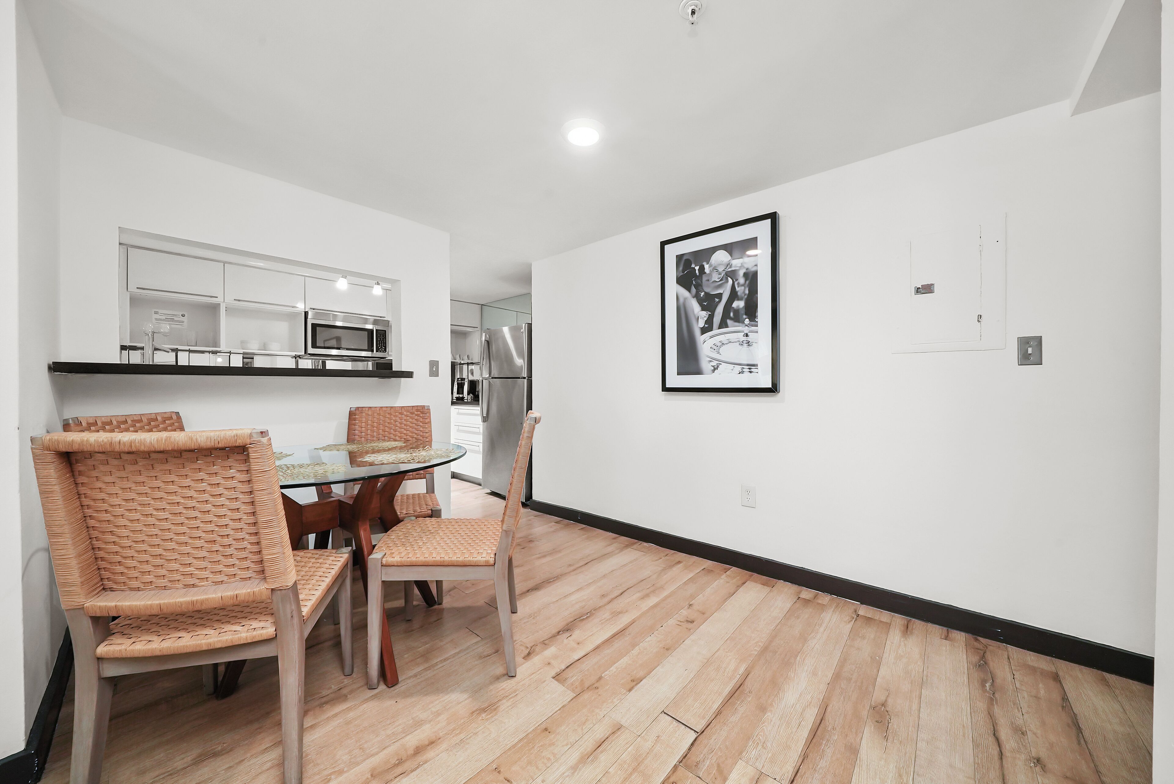 A modern kitchen with white cabinets, a microwave, and a refrigerator. There is a dining area with a glass table and wicker chairs. A framed picture hangs on the wall.