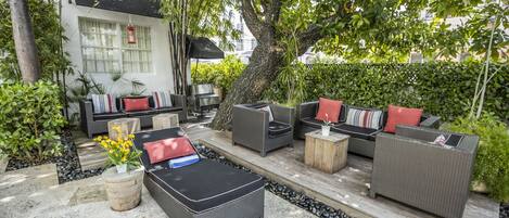 An outdoor patio area with wicker furniture, a large tree, and a view of a building in the background.