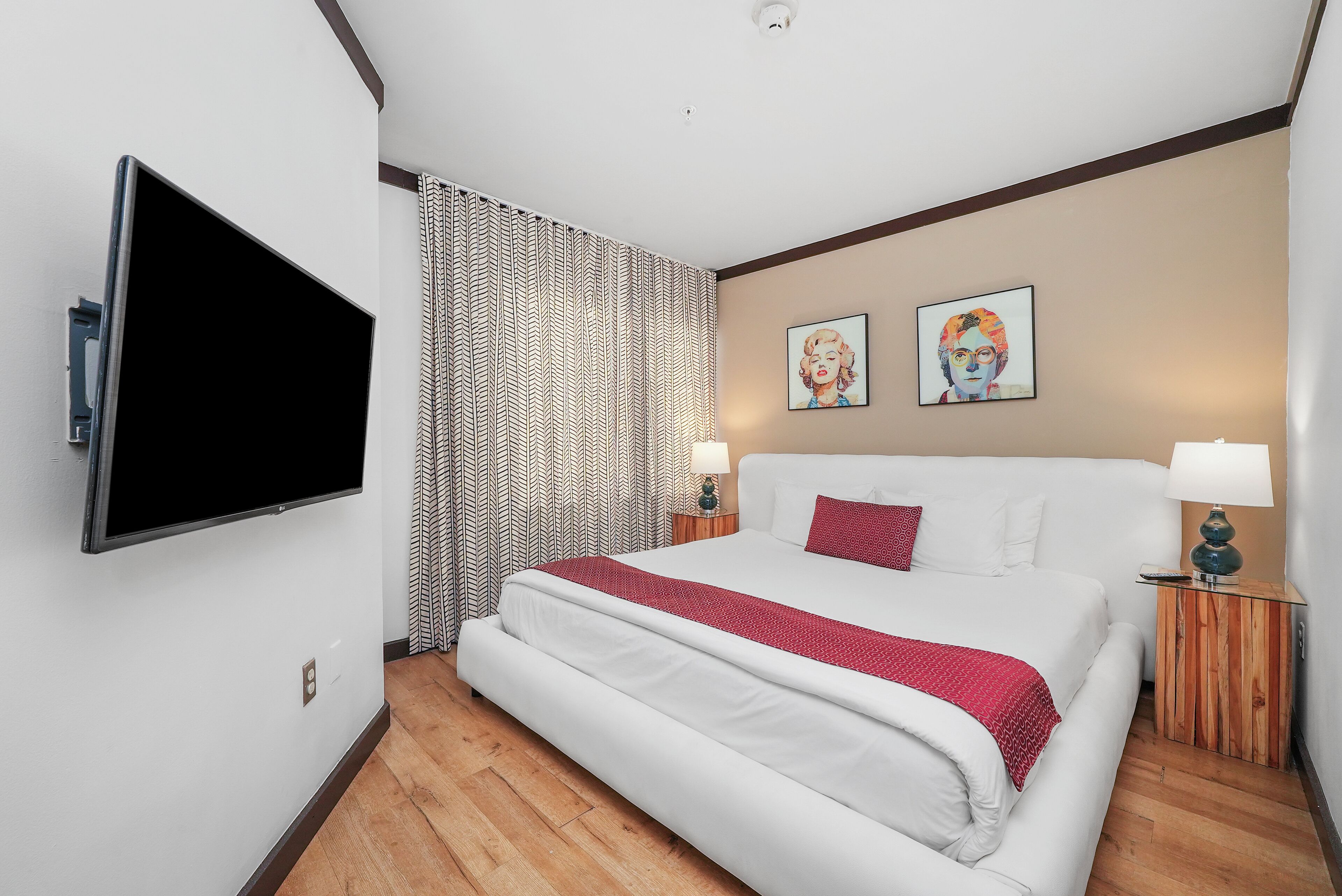 A hotel room with a large flat-screen TV mounted on the wall, a bed with white bedding and a red throw, two bedside tables with lamps, and a patterned curtain.