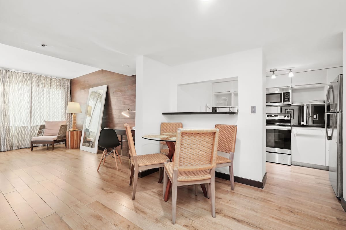 A modern living room with a dining area, a kitchen with stainless steel appliances, and a large window with curtains.
