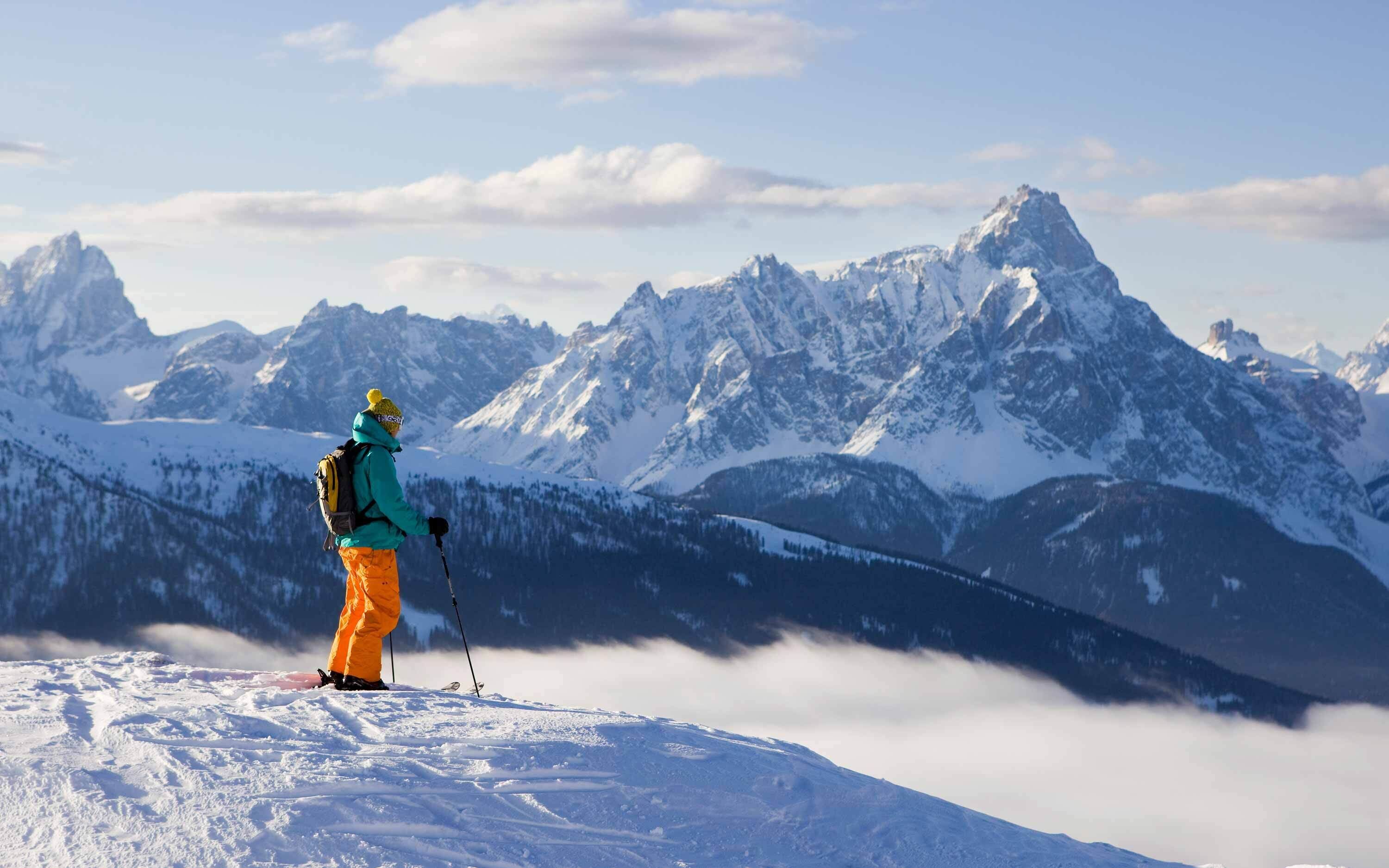 Une personne se tient au sommet d’une montagne enneigée, d’où elle a une vue dégagée sur les montagnes environnantes, également couvertes de neige.