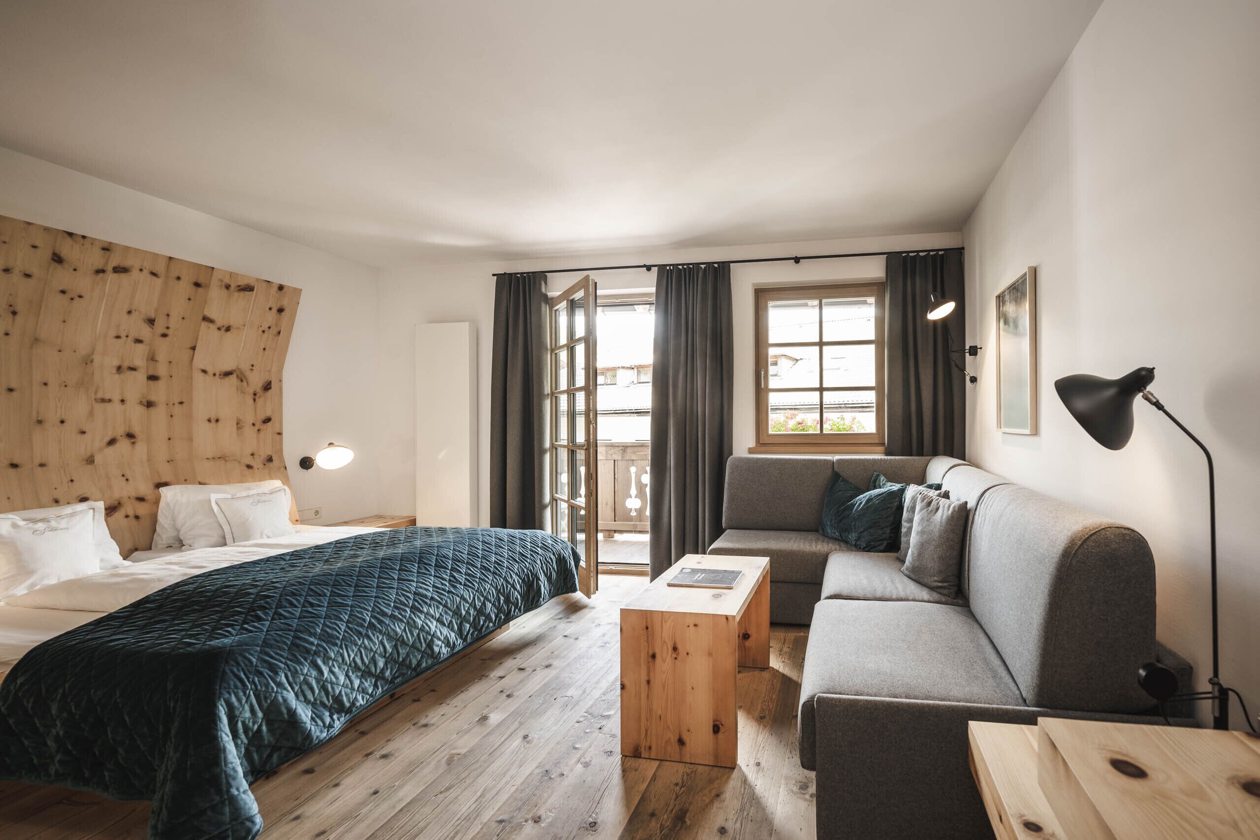 Moderni hotellihuone, jossa on puinen sängynpääty, tummasävyinen peitto, harmaa sohva, puinen sohvapöytä ja suuri ikkuna, jossa on verhot.