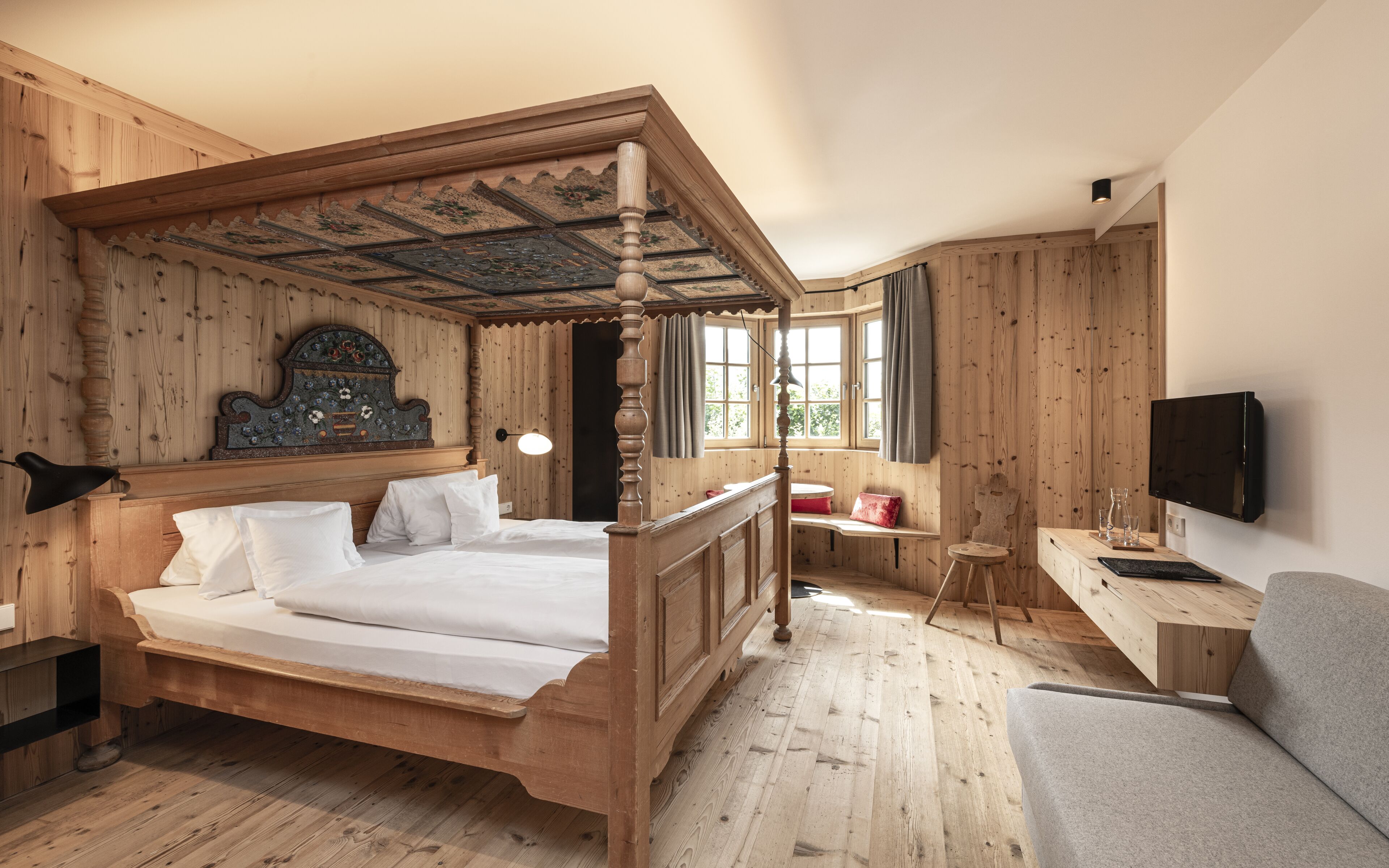 A wooden four-poster bed with a decorative headboard, a desk with a chair, and a television on the wall.