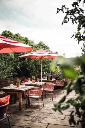 Une terrasse extérieure avec des tables en bois et des chaises en osier, des parasols rouges et des plantes en pot.