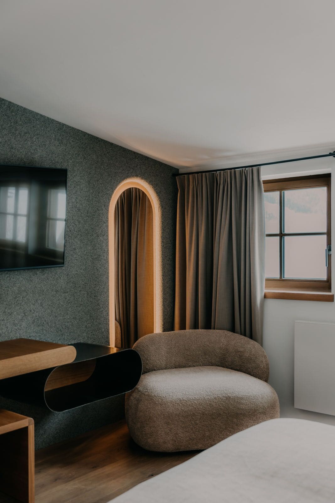 A modern room with a grey textured wall, a flat-screen TV mounted above a wooden desk, a comfortable armchair, and a window with curtains.