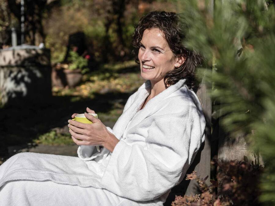 Una persona con bata blanca sentada en un banco al aire libre, sosteniendo una taza amarilla.