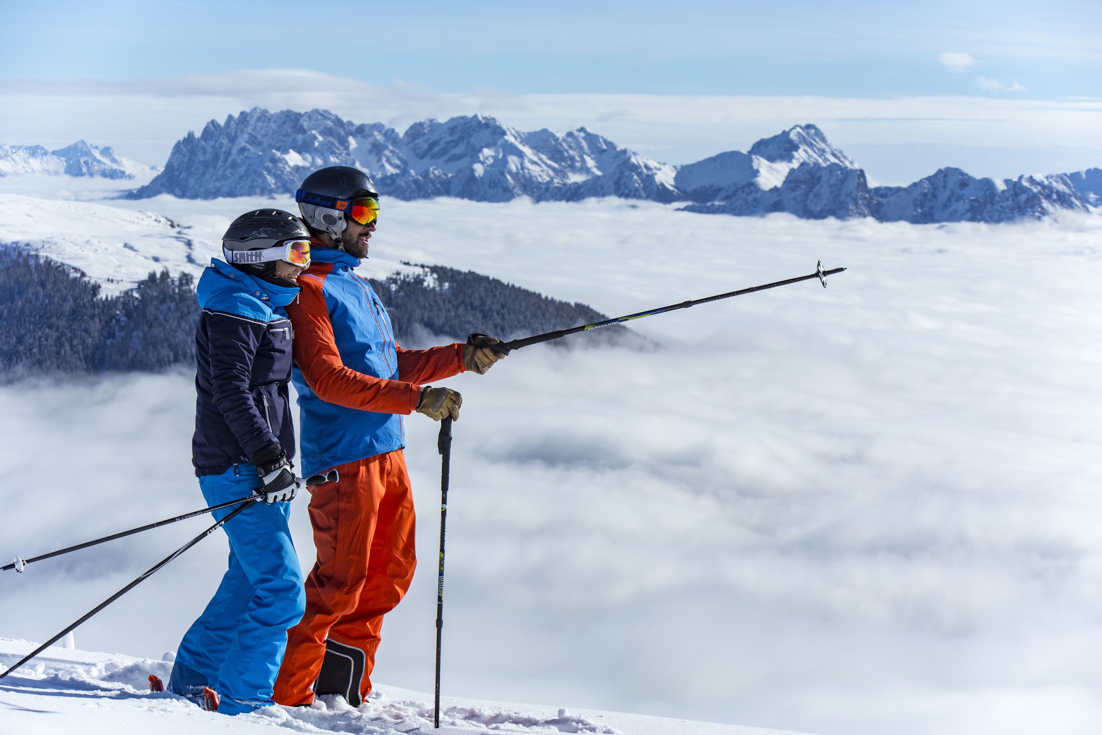 Dos personas con ropa de invierno en una montaña nevada, con vista a picos cubiertos de nieve y nubes abajo.