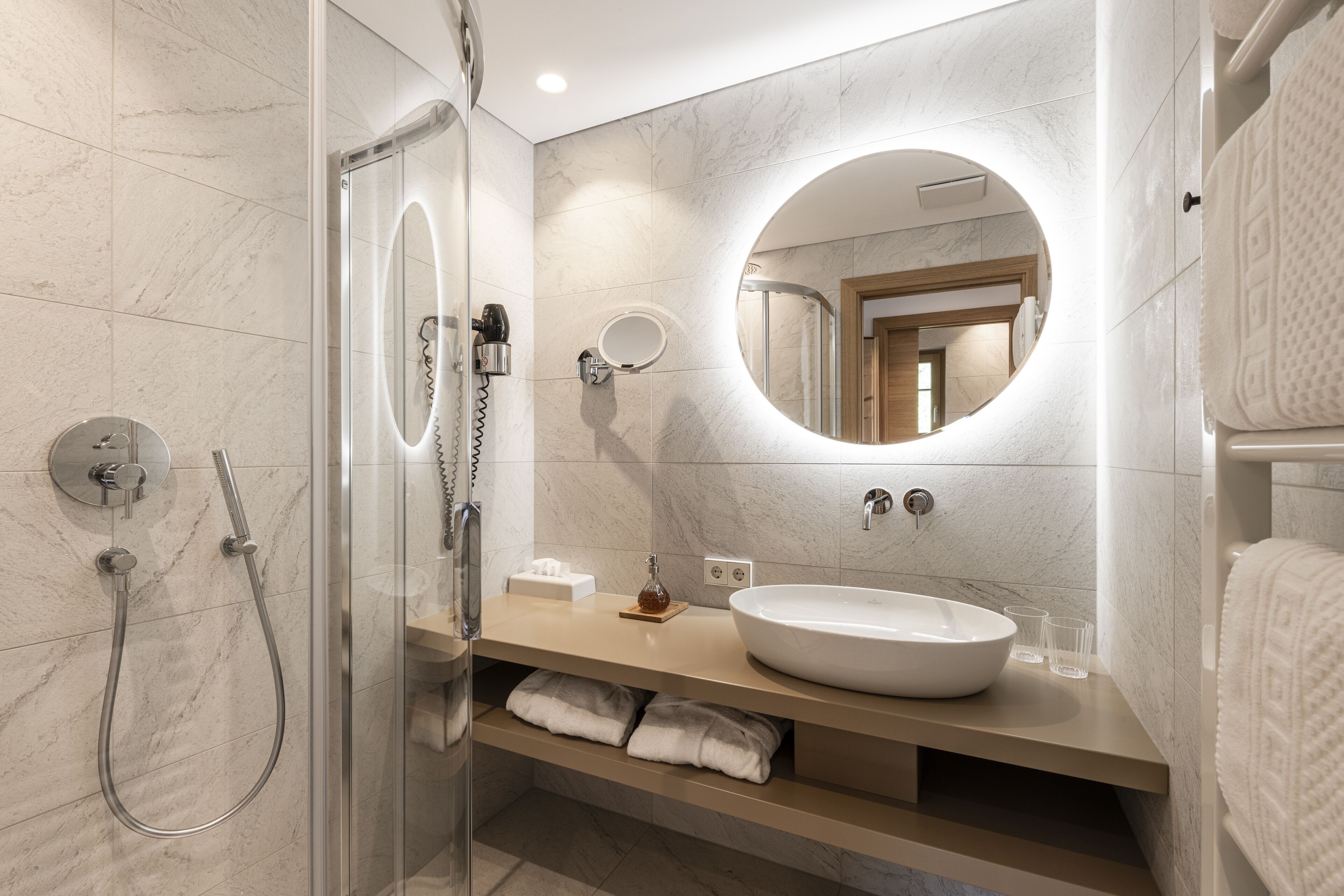 A modern bathroom with a glass-enclosed shower, a round mirror, a beige vanity, and a white sink.