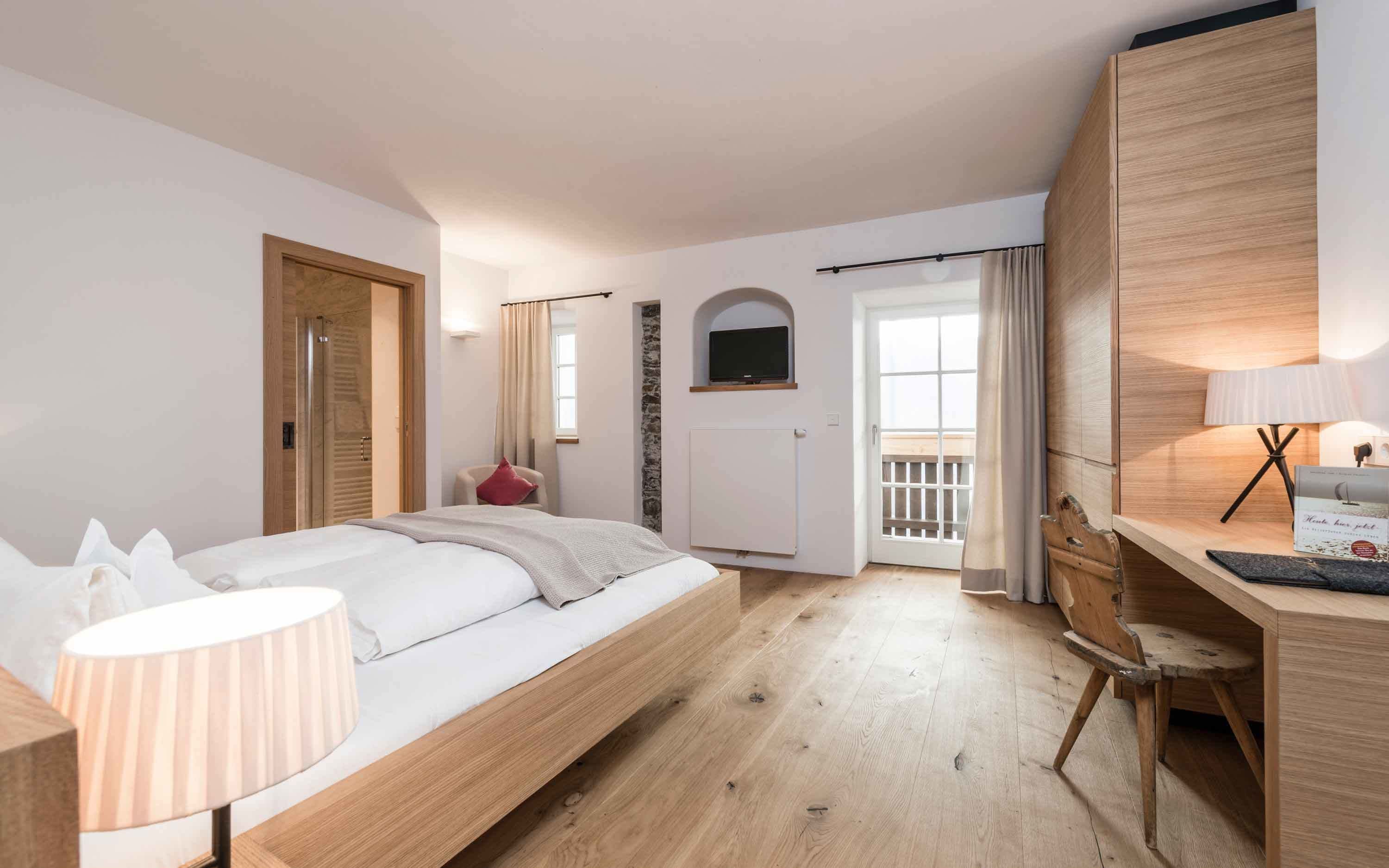 Un dormitorio con cama, mesita de noche, lámpara, escritorio de madera, silla y televisor montado en la pared.