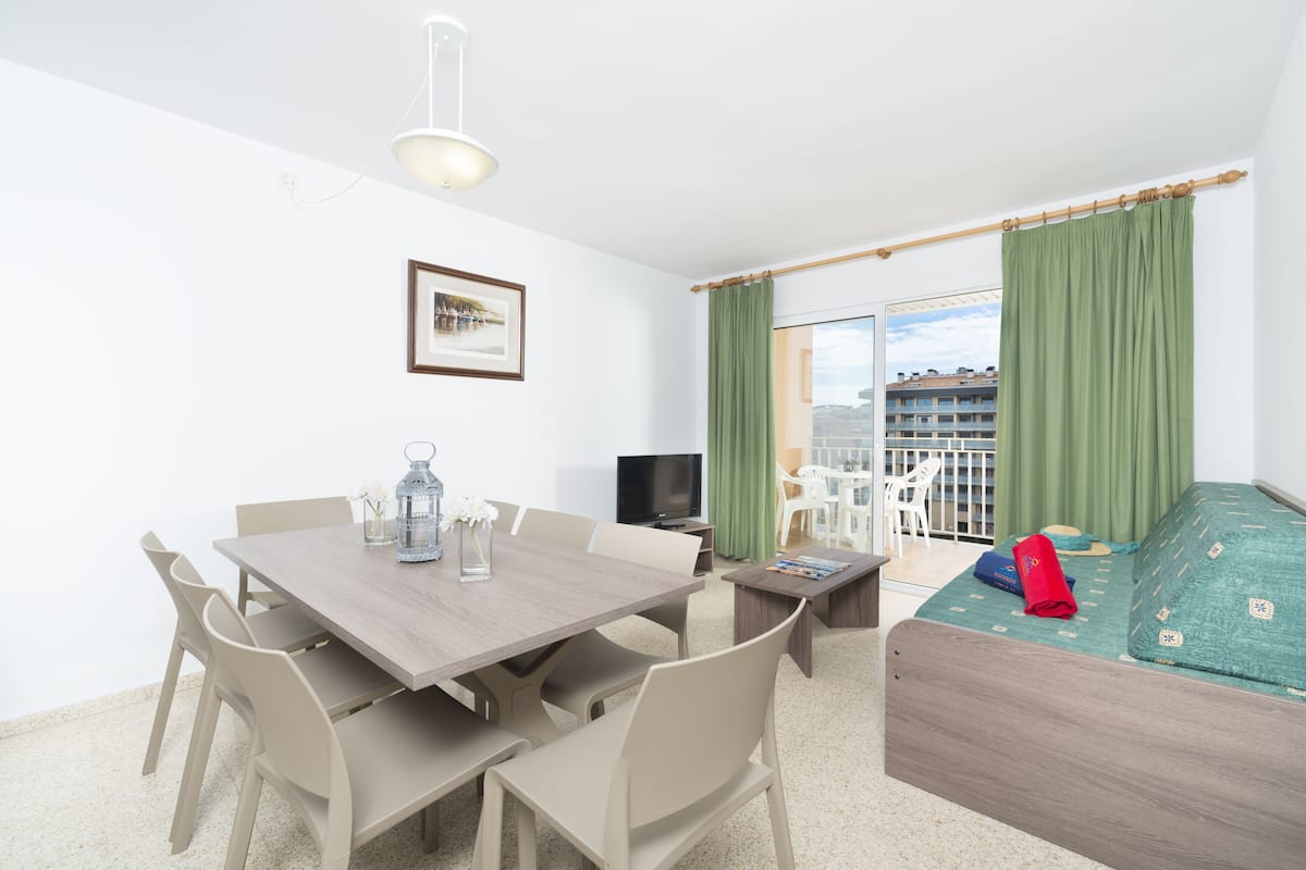 A room with a dining table, chairs, a TV, a bed, and a balcony with curtains.