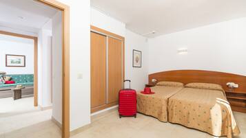 Una habitación de hotel con una cama, una maleta, una mesita de noche, un armario de madera y una pequeña mesa con un sombrero.