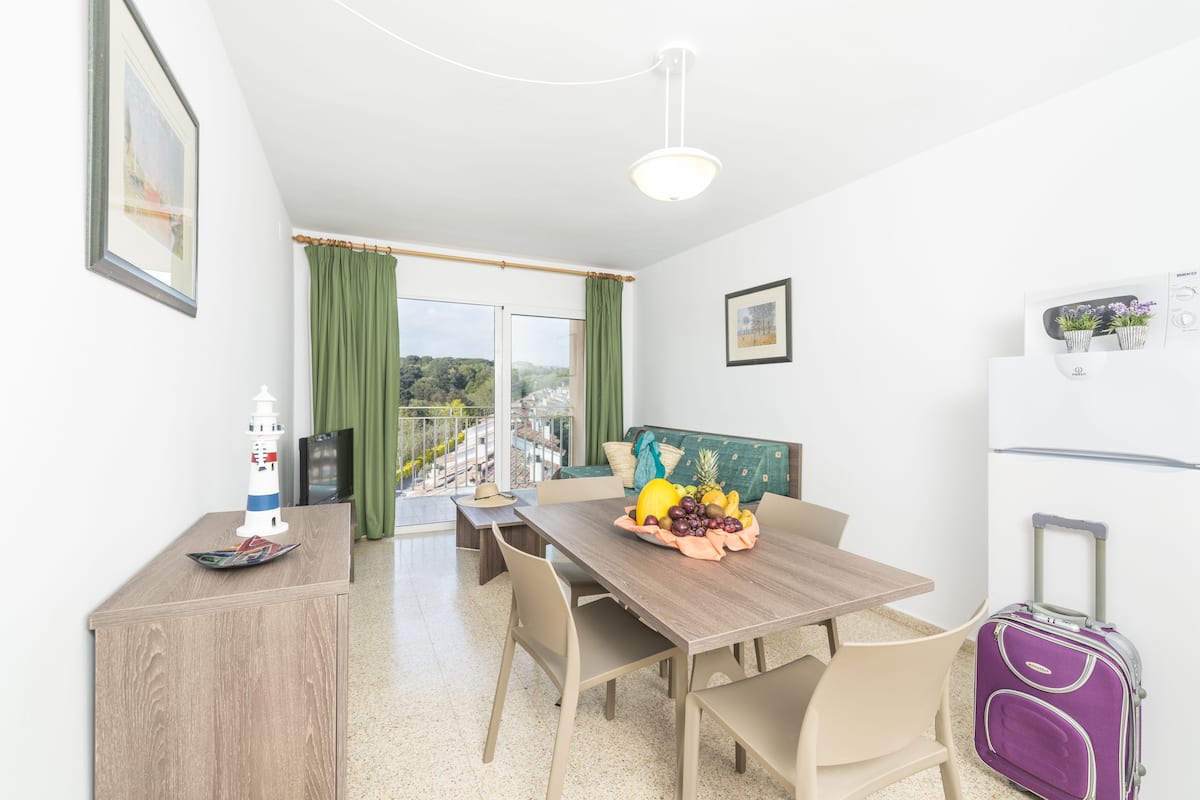 A modern room with a dining table, chairs, a small cabinet, a suitcase, and a view of the outdoors.