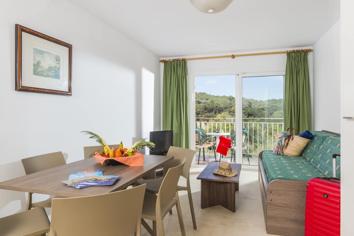 A room with a dining table, chairs, a sofa, a TV, and a balcony with curtains.