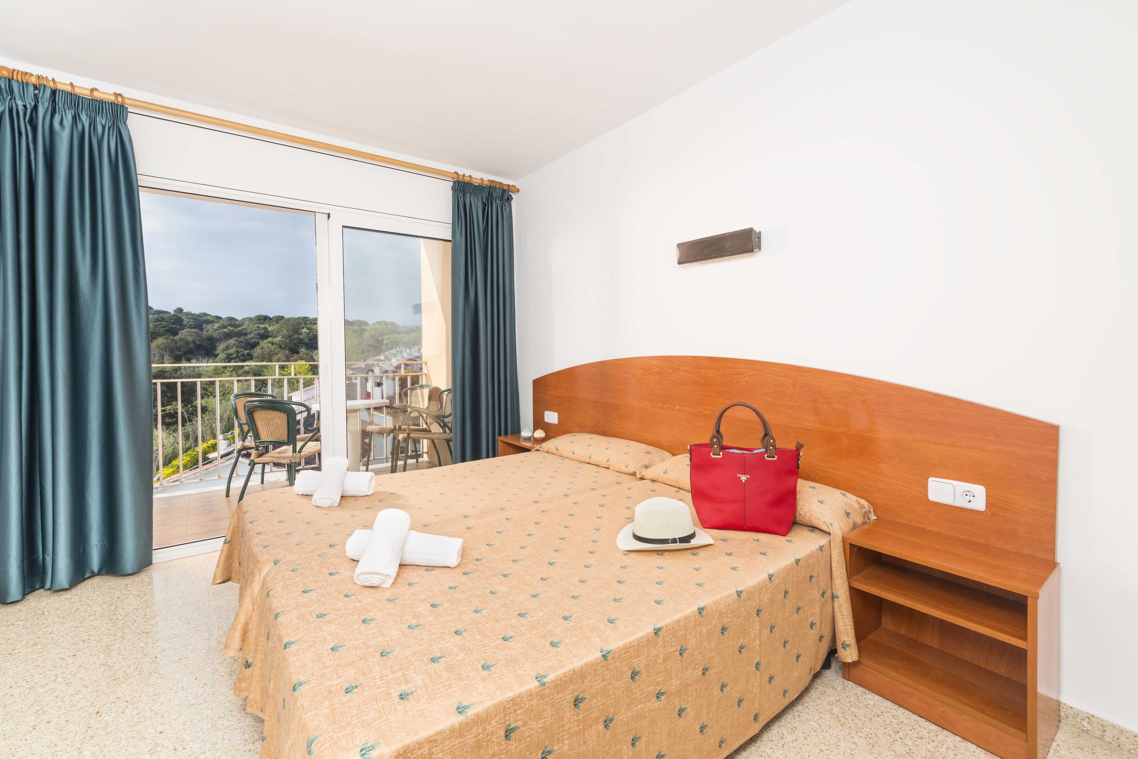 Una habitación de hotel con una cama, una mesita de noche, un bolso rojo, un sombrero y vistas a un balcón con sillas y una mesa.