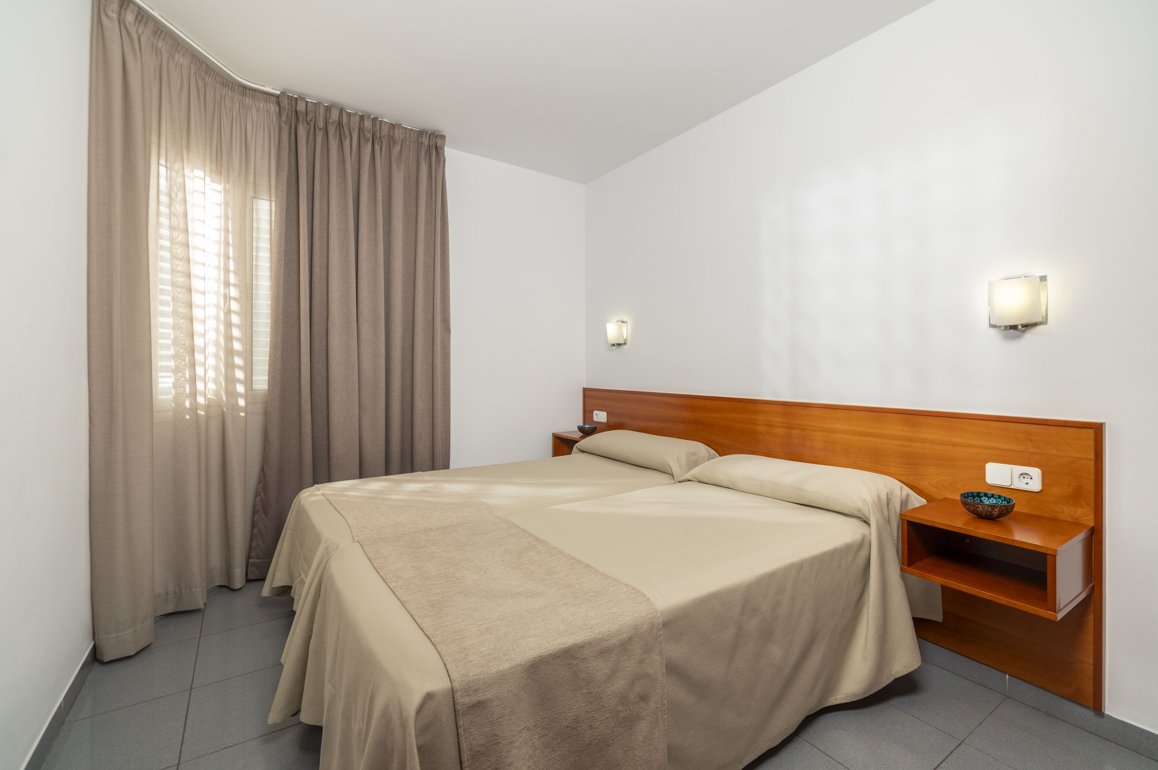 A neatly made bed with a wooden headboard, a bedside table with a bowl, and a wall-mounted light fixture.