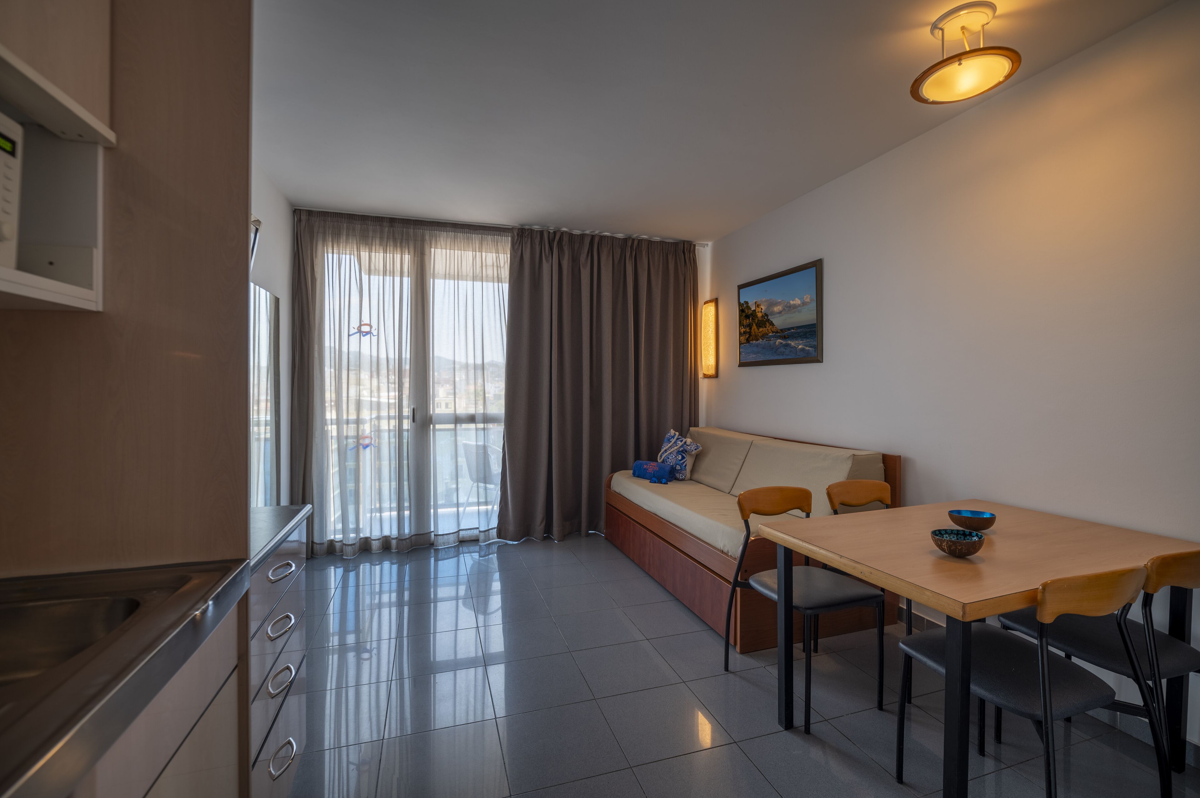 Ein modernes Hotelzimmer mit einer Küchenzeile, einem Essbereich mit Tisch und Stühlen, einer Sitzecke und einem großen Fenster mit Vorhängen.
