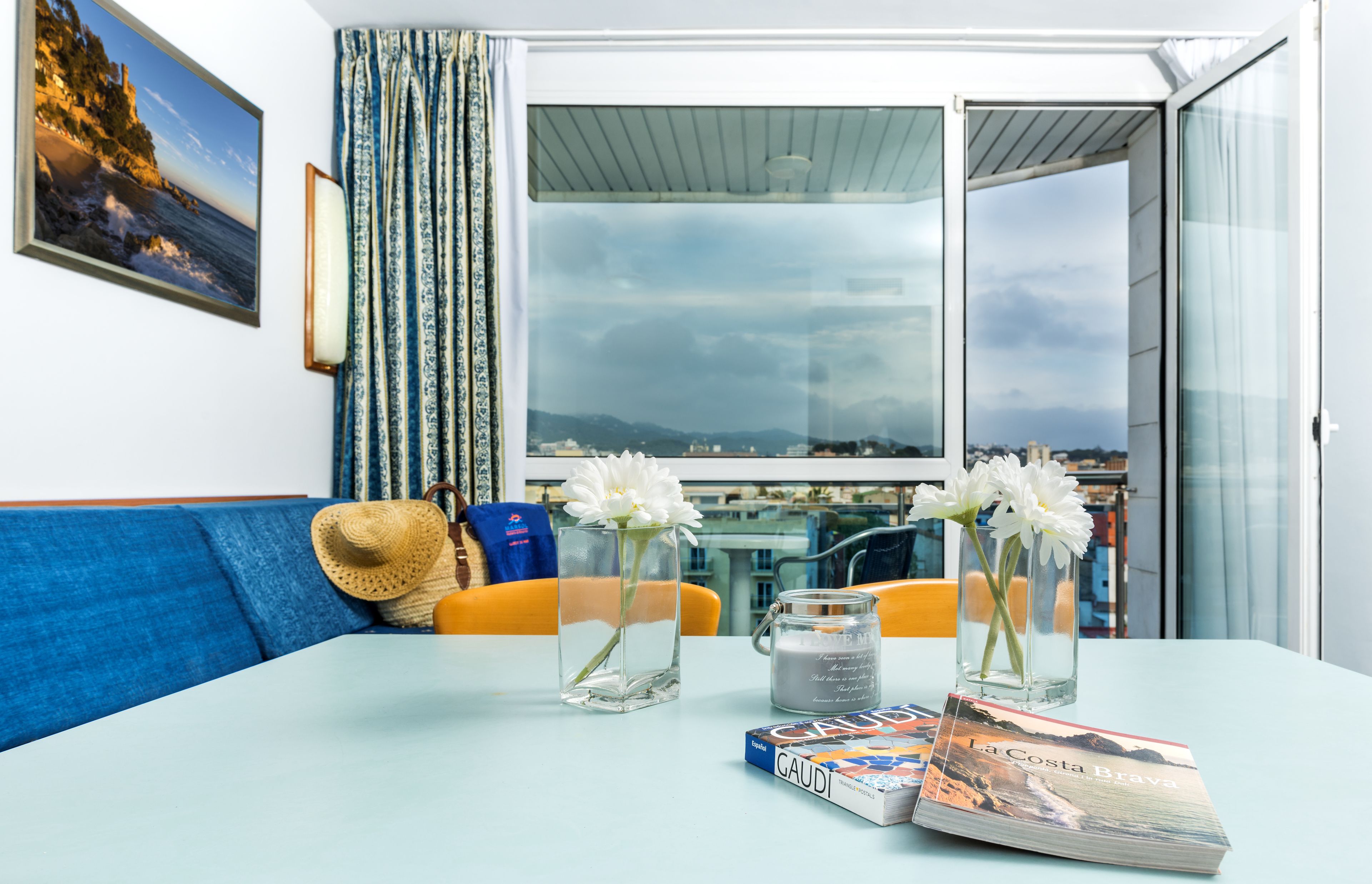 A room with a view of the city, a table with a vase of flowers, a candle, and a book titled "La Costa Brava".