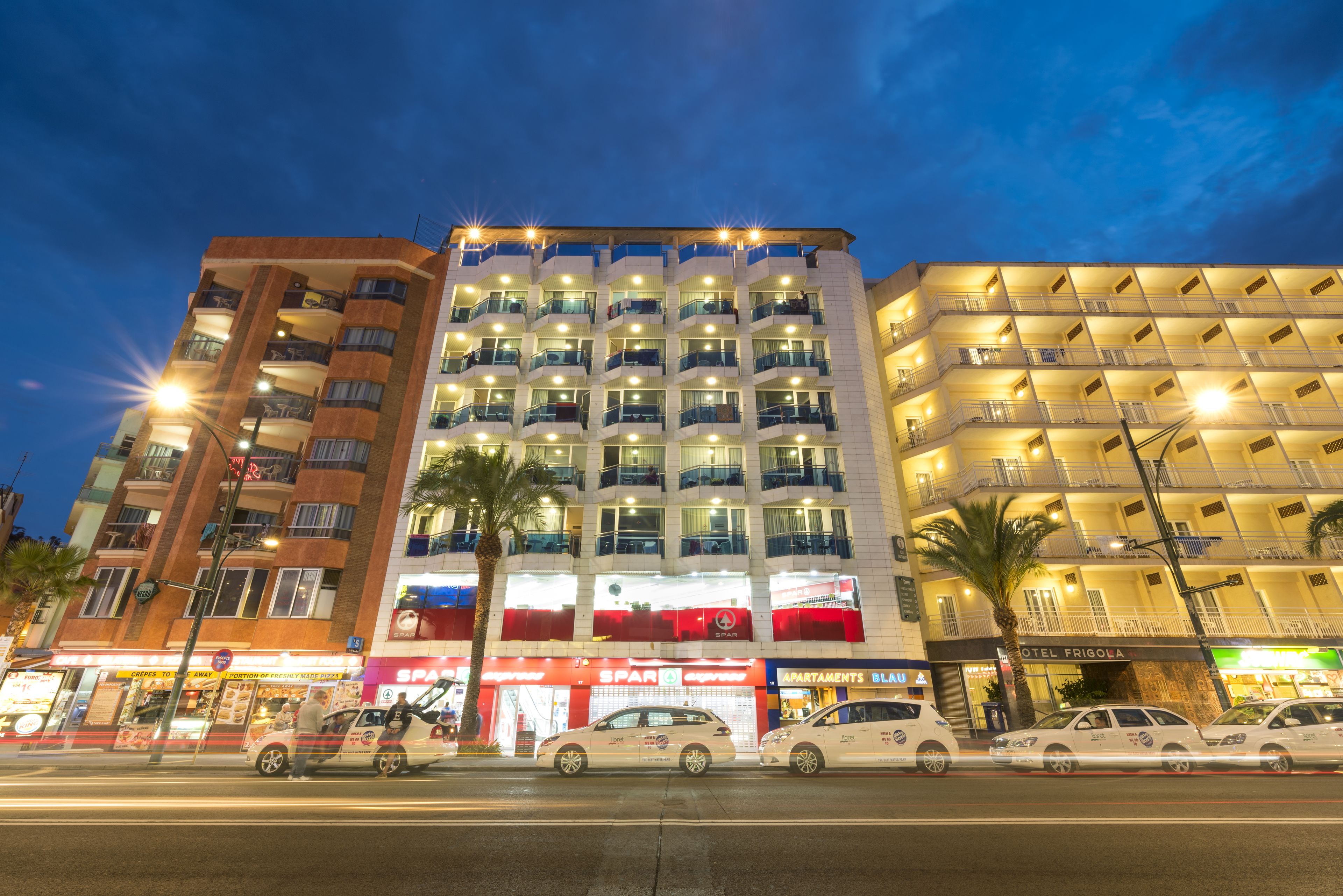 Un hotel de varios pisos con balcones, un restaurante a la vista y coches aparcados.