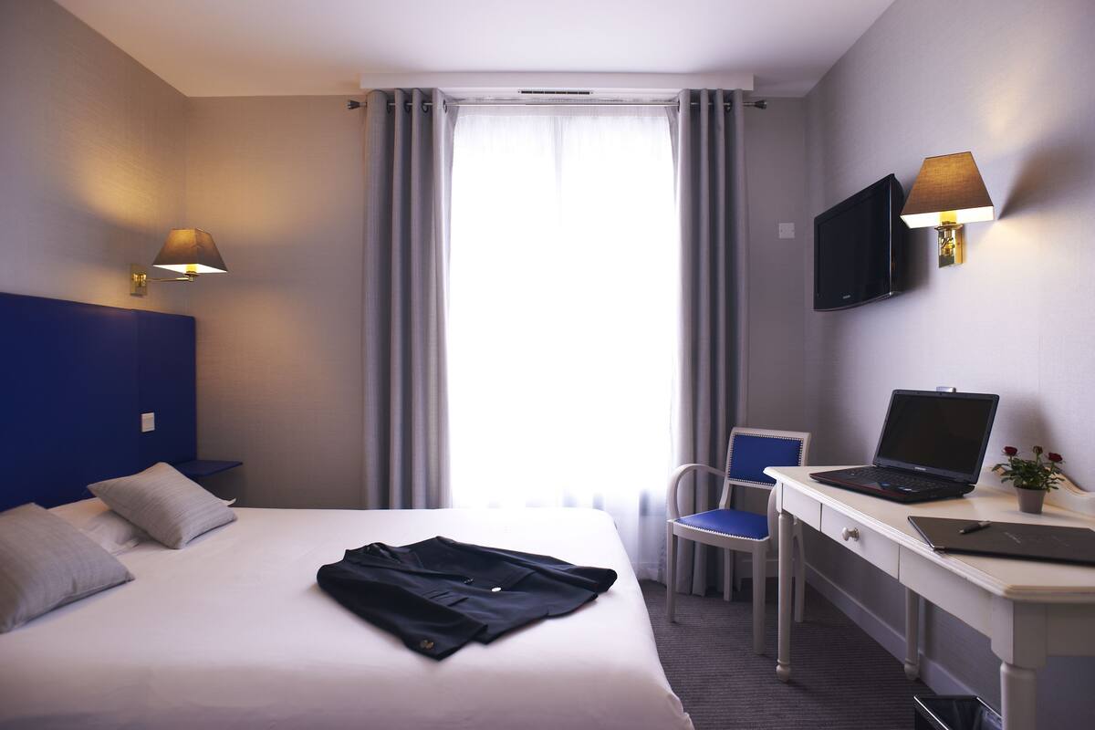 Ein Hotelzimmer mit Bett, Schreibtisch mit Laptop, Stuhl, Fernseher und einem Fenster mit Vorhängen.