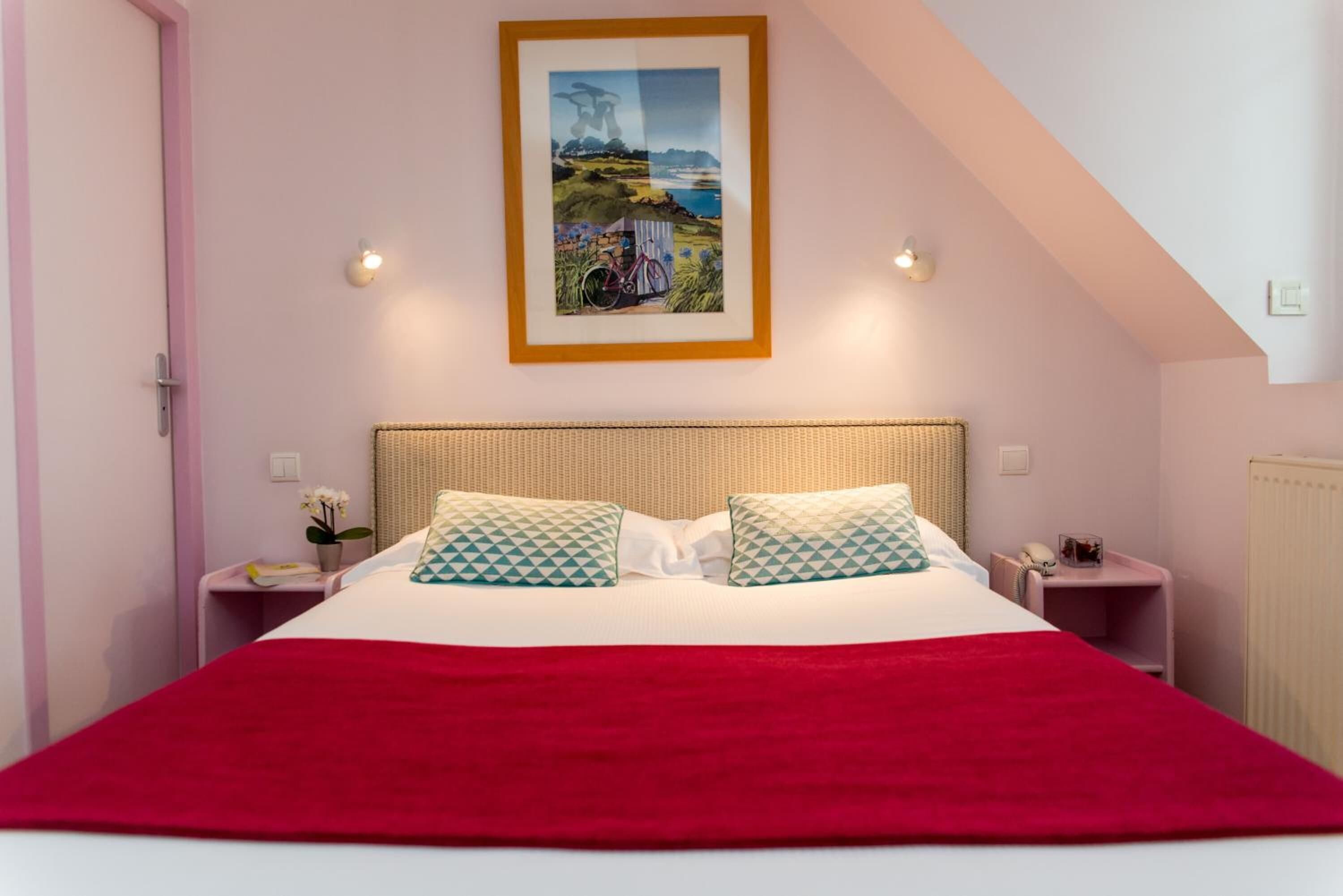 Une chambre à coucher avec un grand lit, une couverture rouge, deux coussins à motifs, un tableau représentant un paysage encadré et deux tables de chevet avec des lampes.