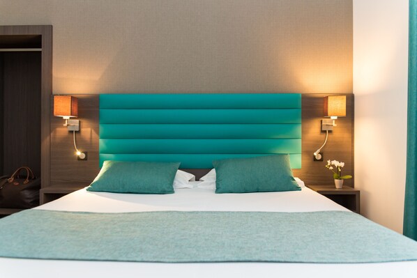 Una cama bien hecha con ropa de cama color turquesa, dos almohadas a juego y una manta color turquesa. Hay dos mesitas de noche con lámparas y un pequeño jarrón con flores.