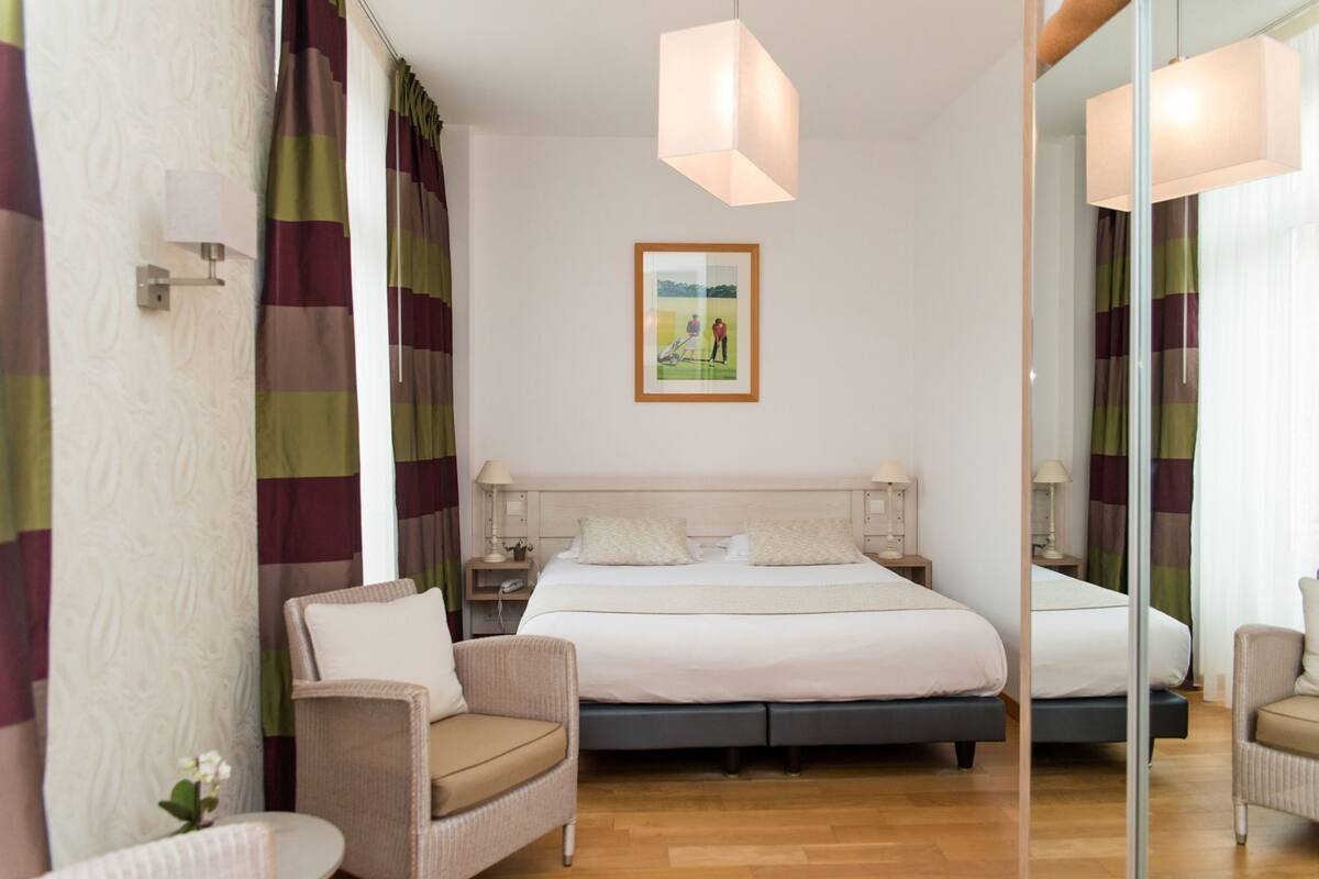 Ein Hotelzimmer mit Bett, zwei Nachttischen, einem Sessel und einem Spiegel, sowie einem Bild an der Wand.